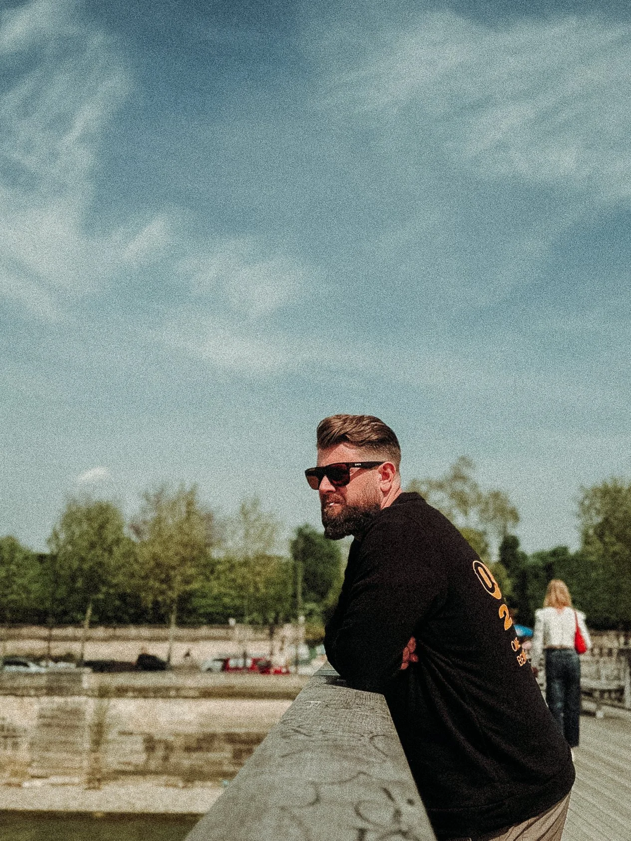 Man with sunglasses leaning on a wooden railing, looking out over a scenic view with trees and a river, under a cloudy sky.