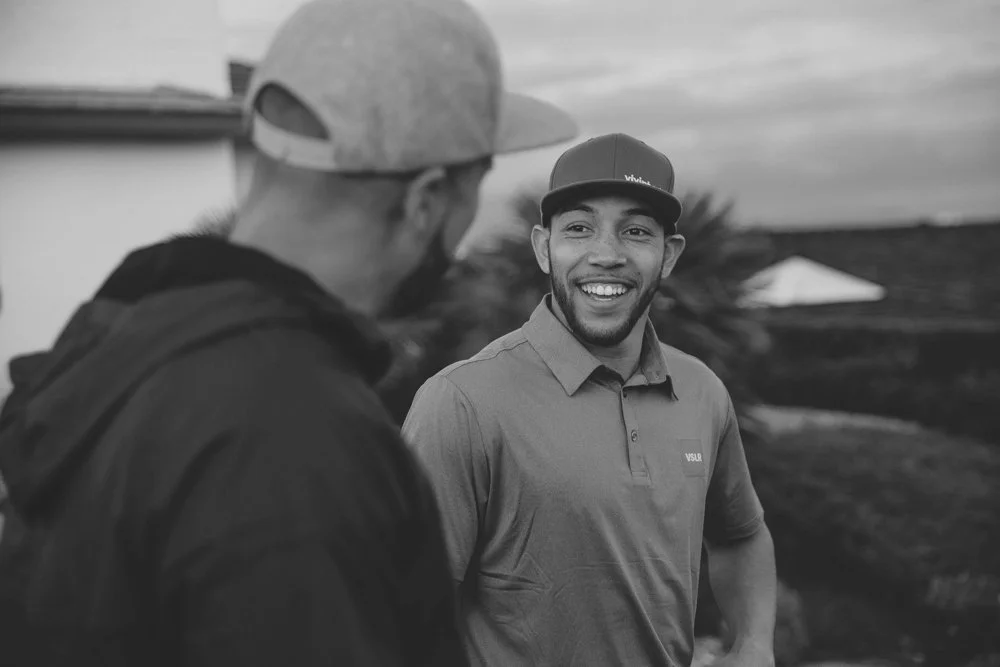 Two men wearing baseball caps having a friendly conversation outdoors, one smiling and looking towards the other.