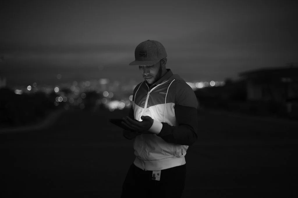 Man in a hoodie and cap looking at a smartphone at night with city lights in the background.