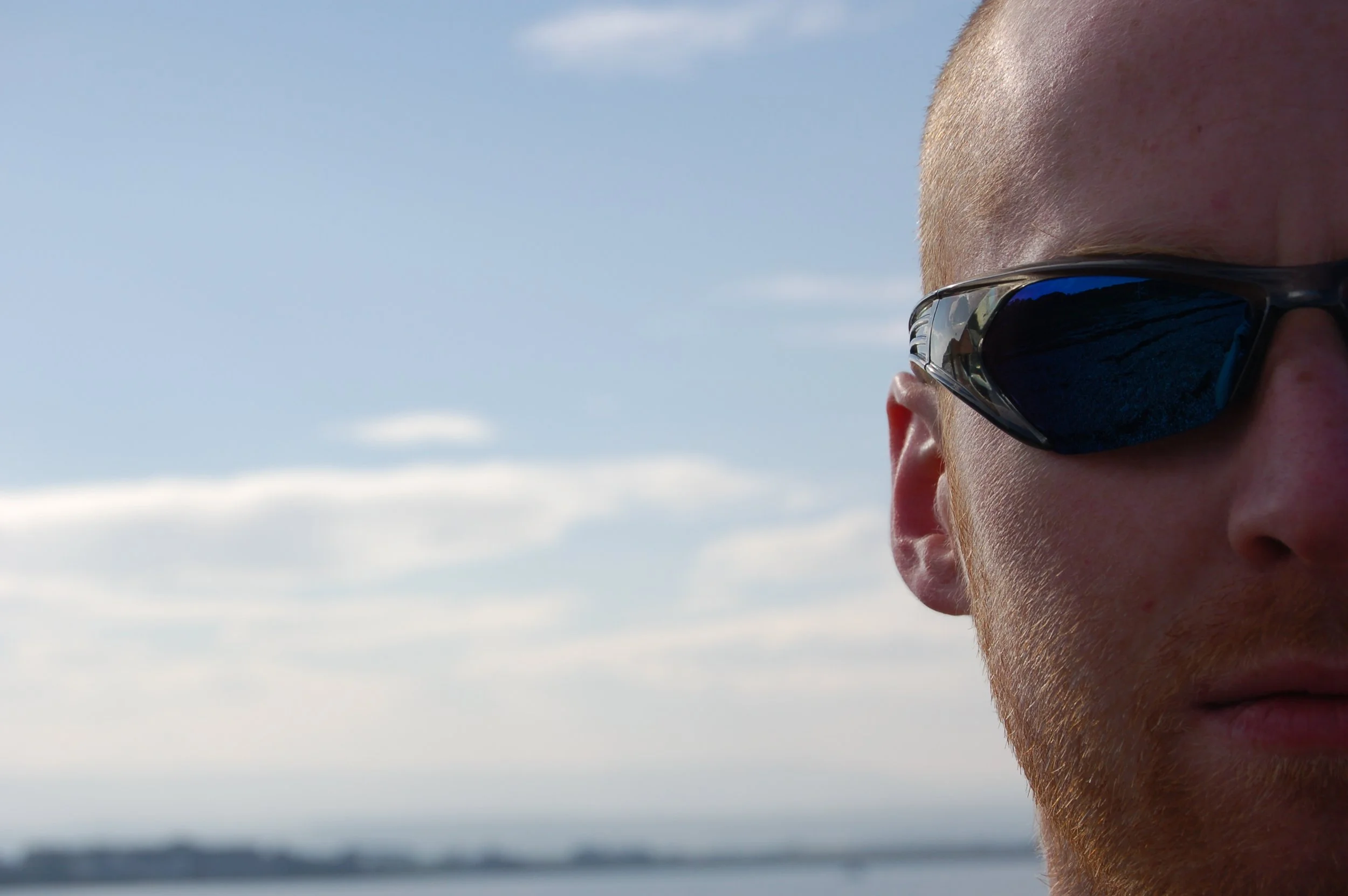 Close-up of a man with red beard wearing black sports sunglasses, outdoors near water with a sky background.