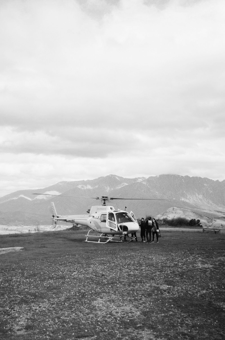 A helicopter parked on a grassy area with four people standing beside it, mountains in the background, under cloudy skies.