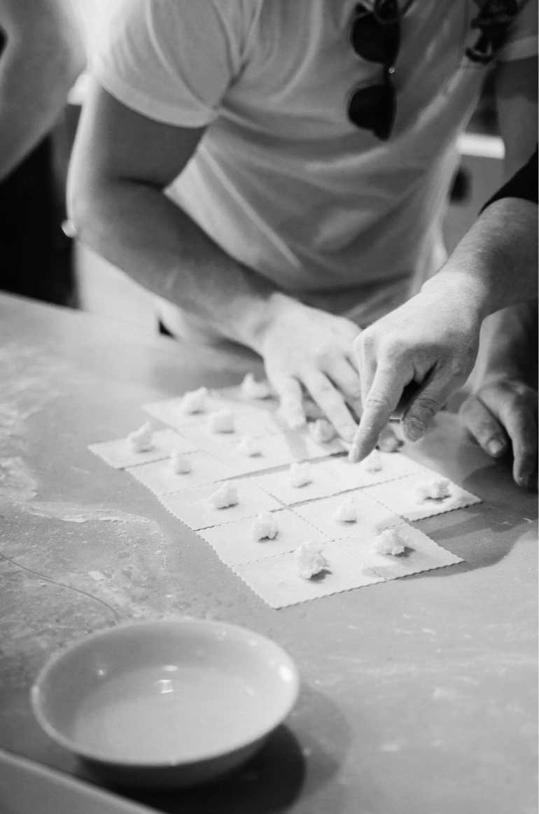 People making dumplings or similar food on a counter, with small portions of filling placed on pieces of dough, in a kitchen setting.