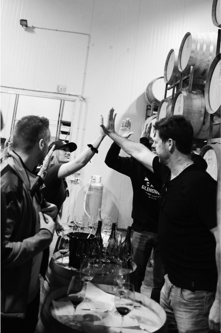 People giving high-fives at a wine tasting event with wine glasses and bottles on a table in the foreground, and wine barrels on the wall in the background.