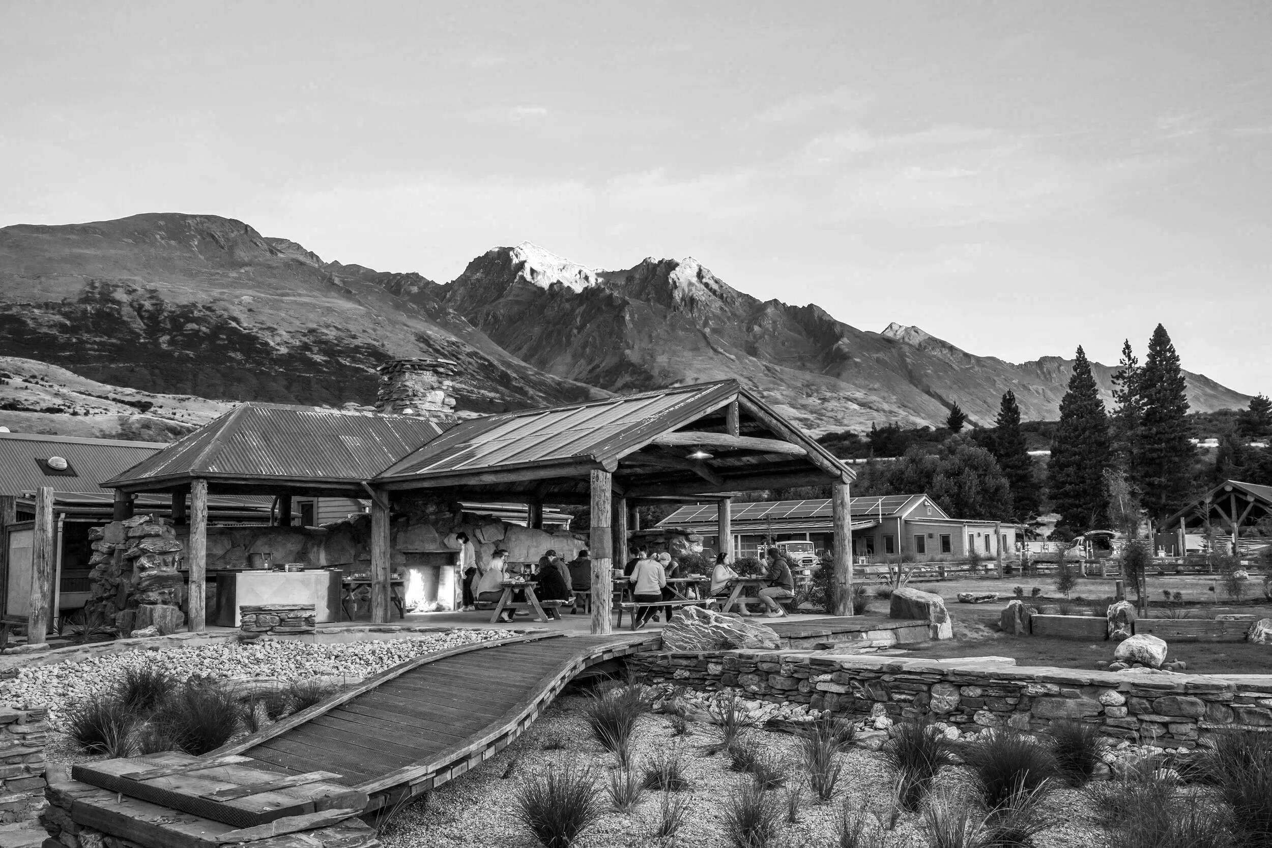 A black and white photo of a mountain landscape with a rustic outdoor dining area in the foreground, featuring a covered pavilion and people sitting at picnic tables.