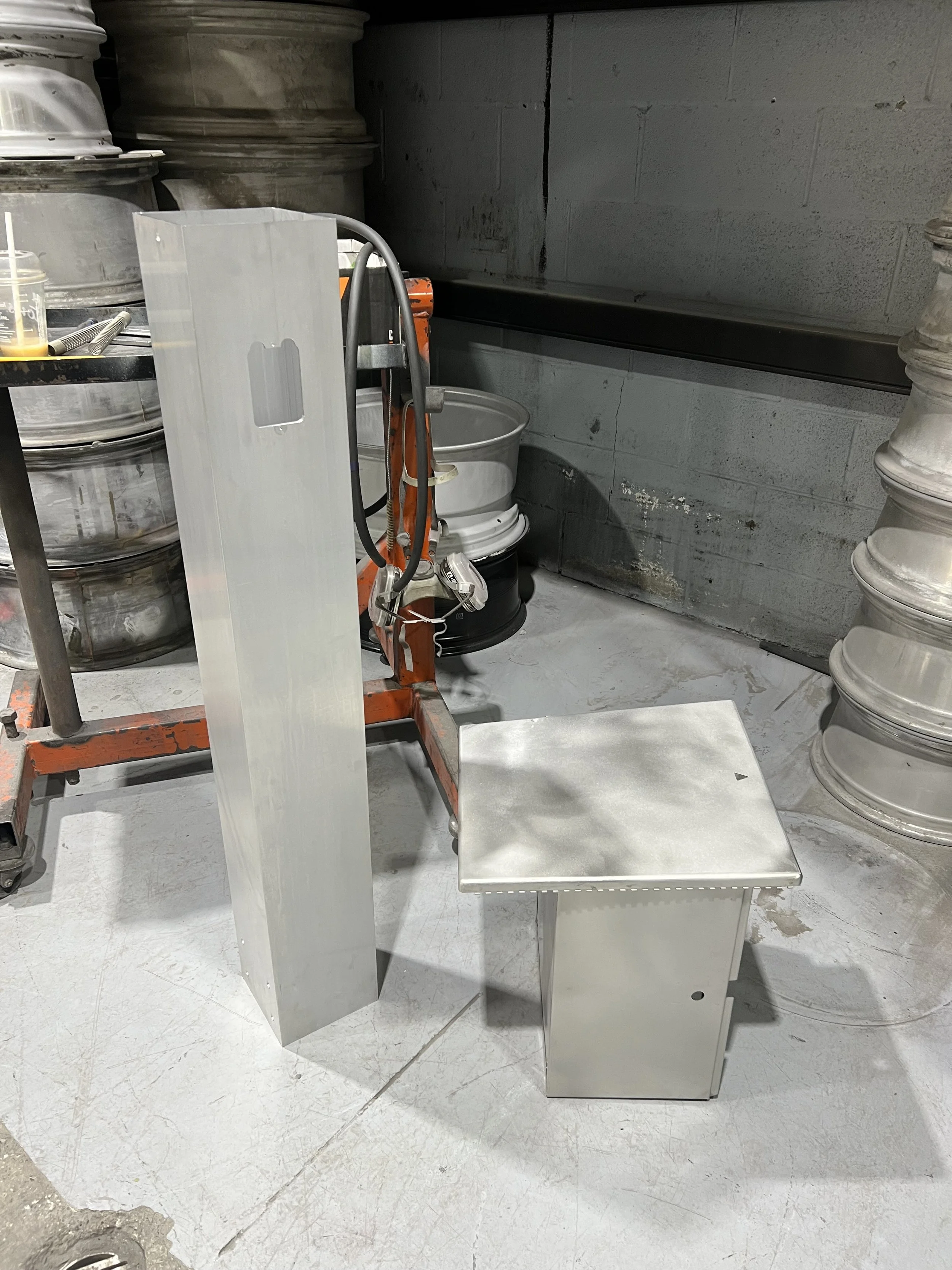 Metal stand with a tall rectangular panel and a small square table with a door, set in a workshop with concrete walls and metal containers.