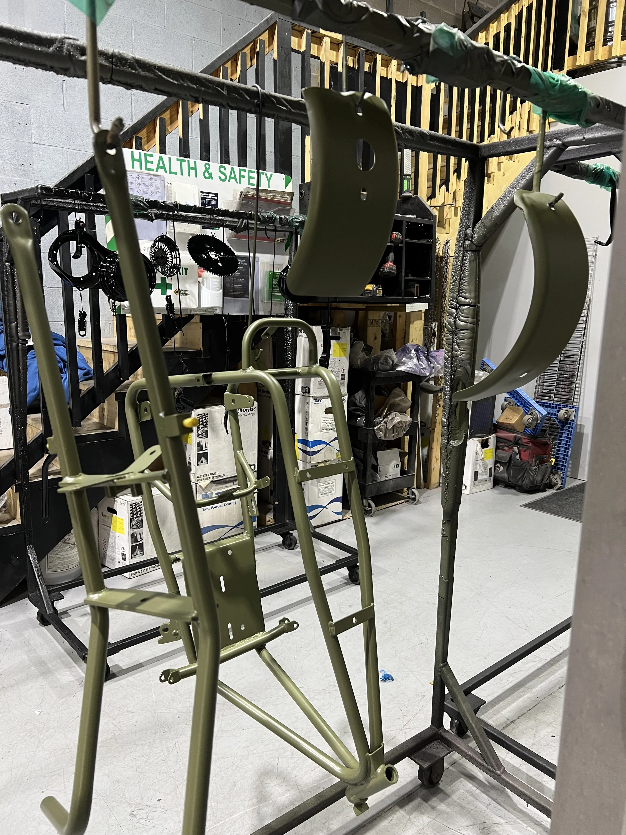 Disassembled military-style green walker and chair hanging on a rack inside a workshop or storage area with tools and supplies in the background.