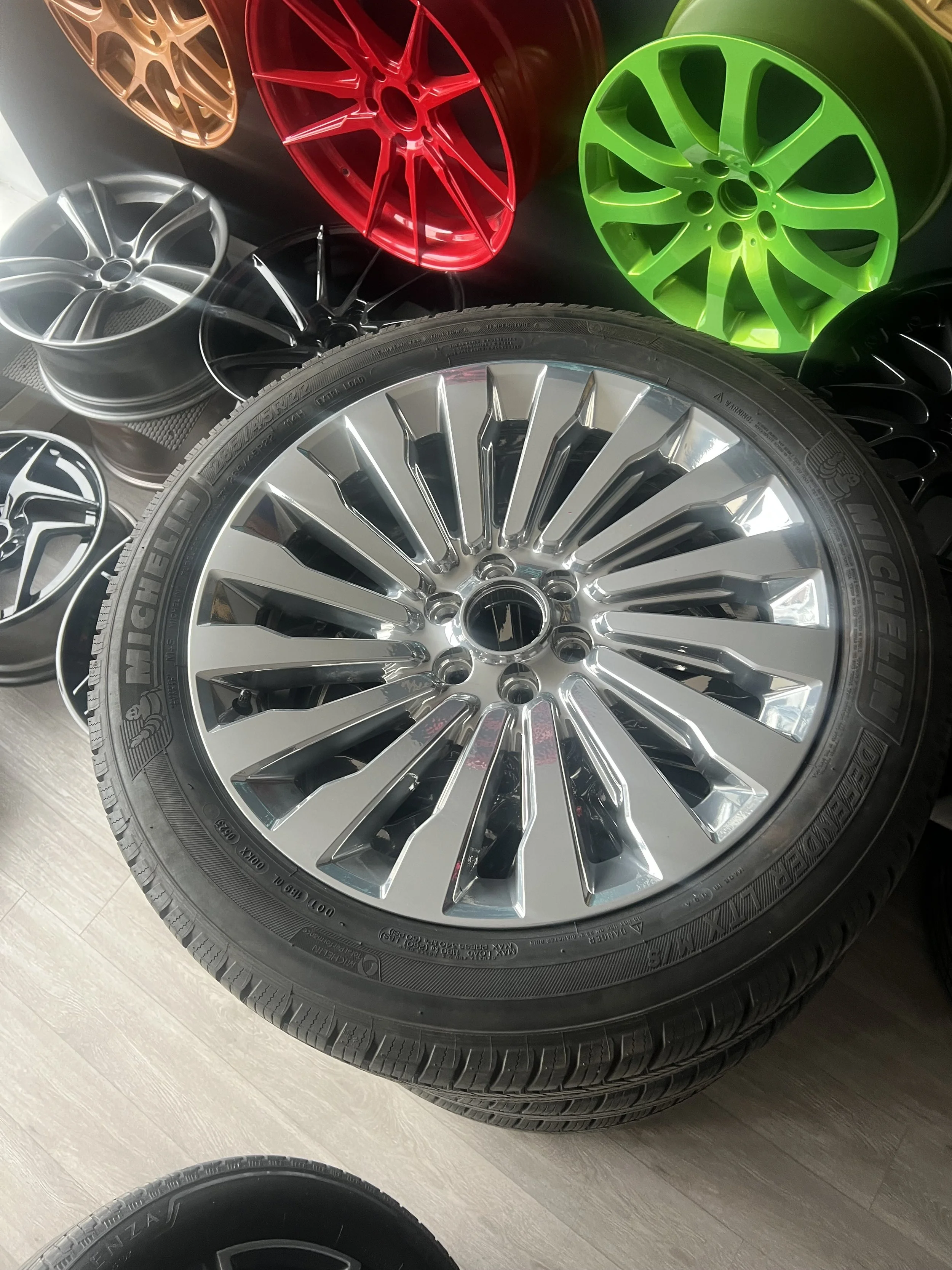 A stack of shiny, multi-spoke alloy wheels with a tire, displayed among colorful wheels including red, green, black, and bronze.