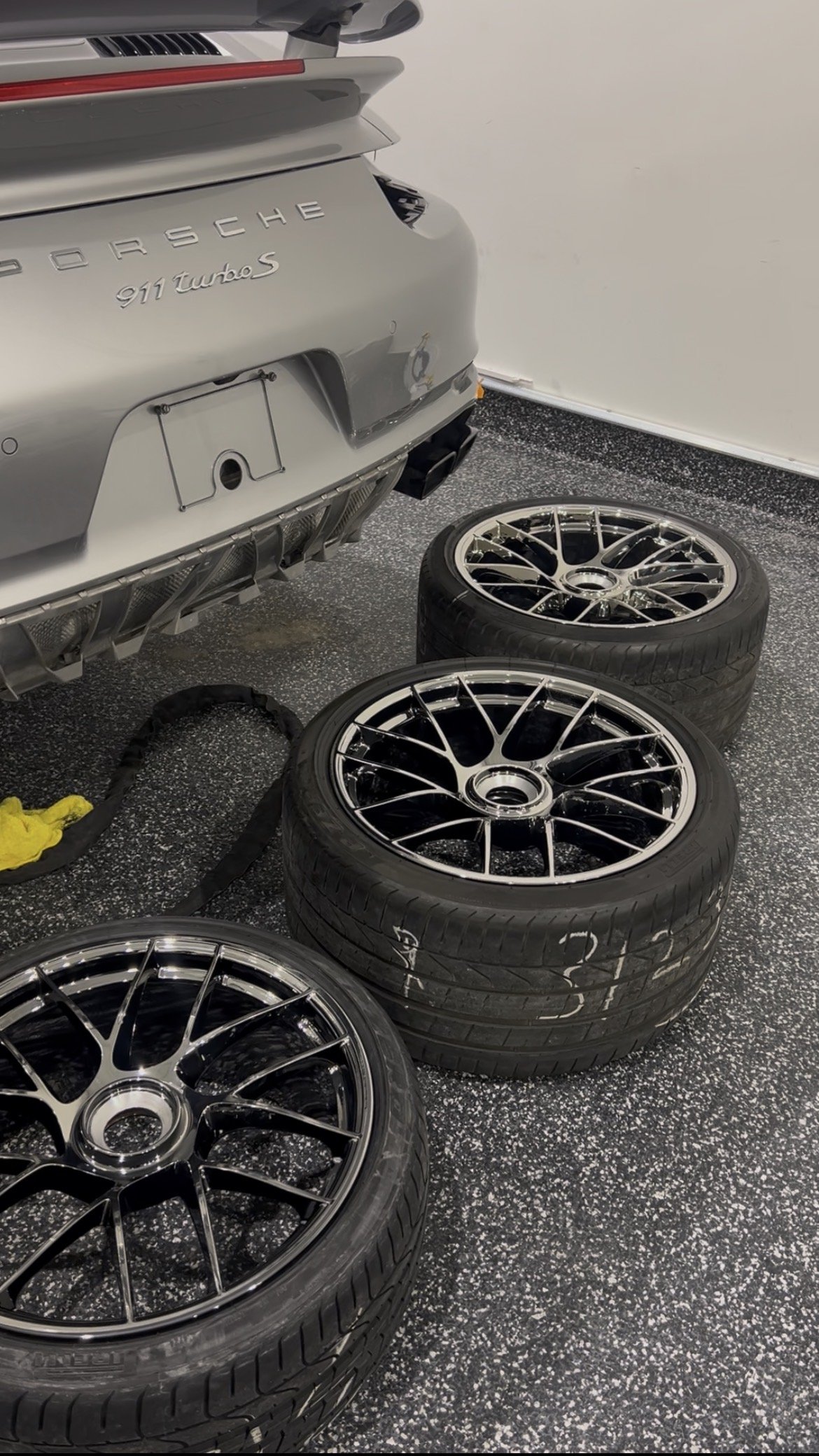 A silver Porsche 911 Turbo S with three black tire rims and tires arranged on the floor nearby in a garage.