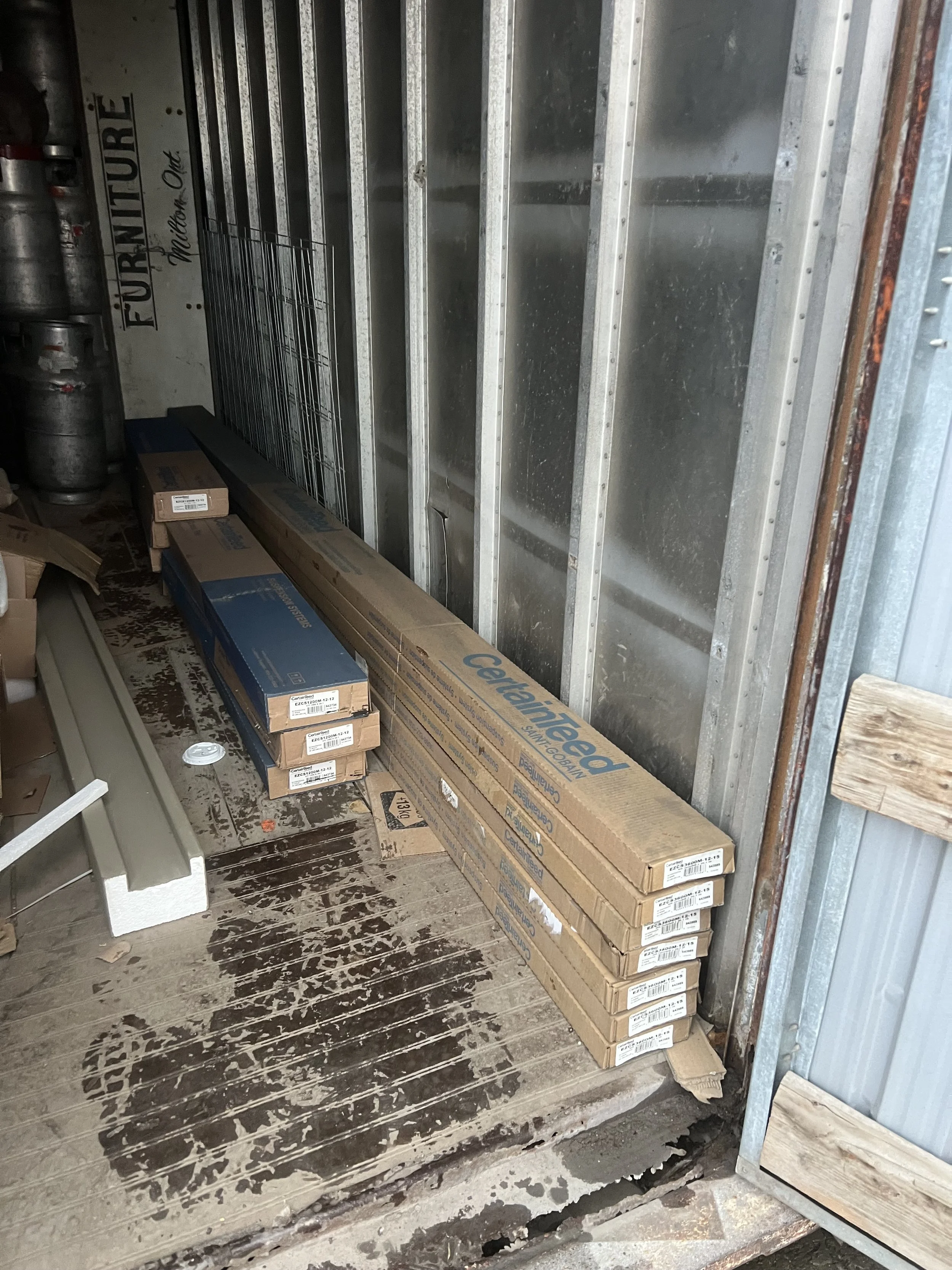 Stacked boxes of construction materials inside a storage unit with metal framing and wooden floor.