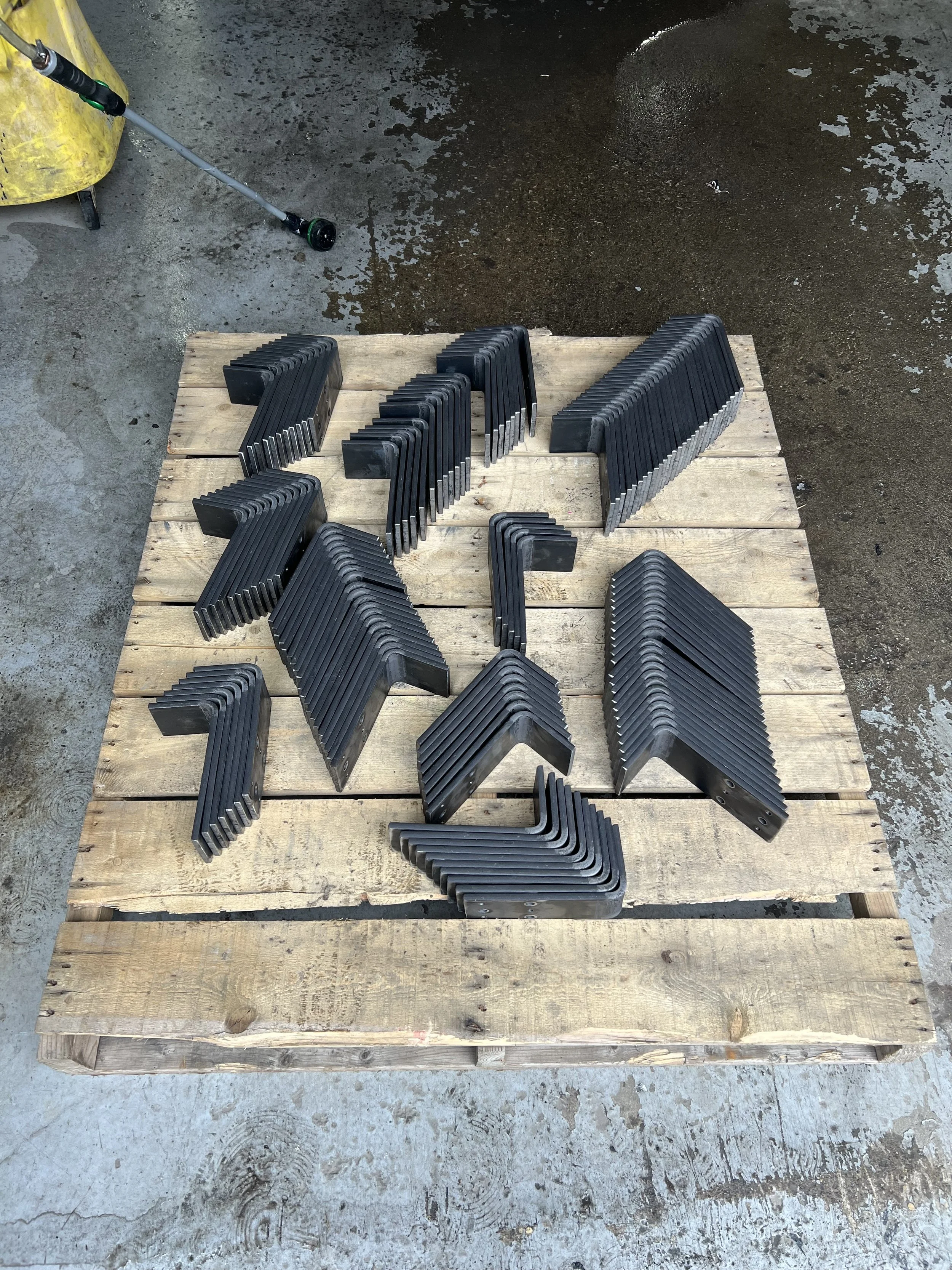 Multiple black L-shaped and U-shaped metal brackets arranged on a wooden pallet in an industrial or construction setting.