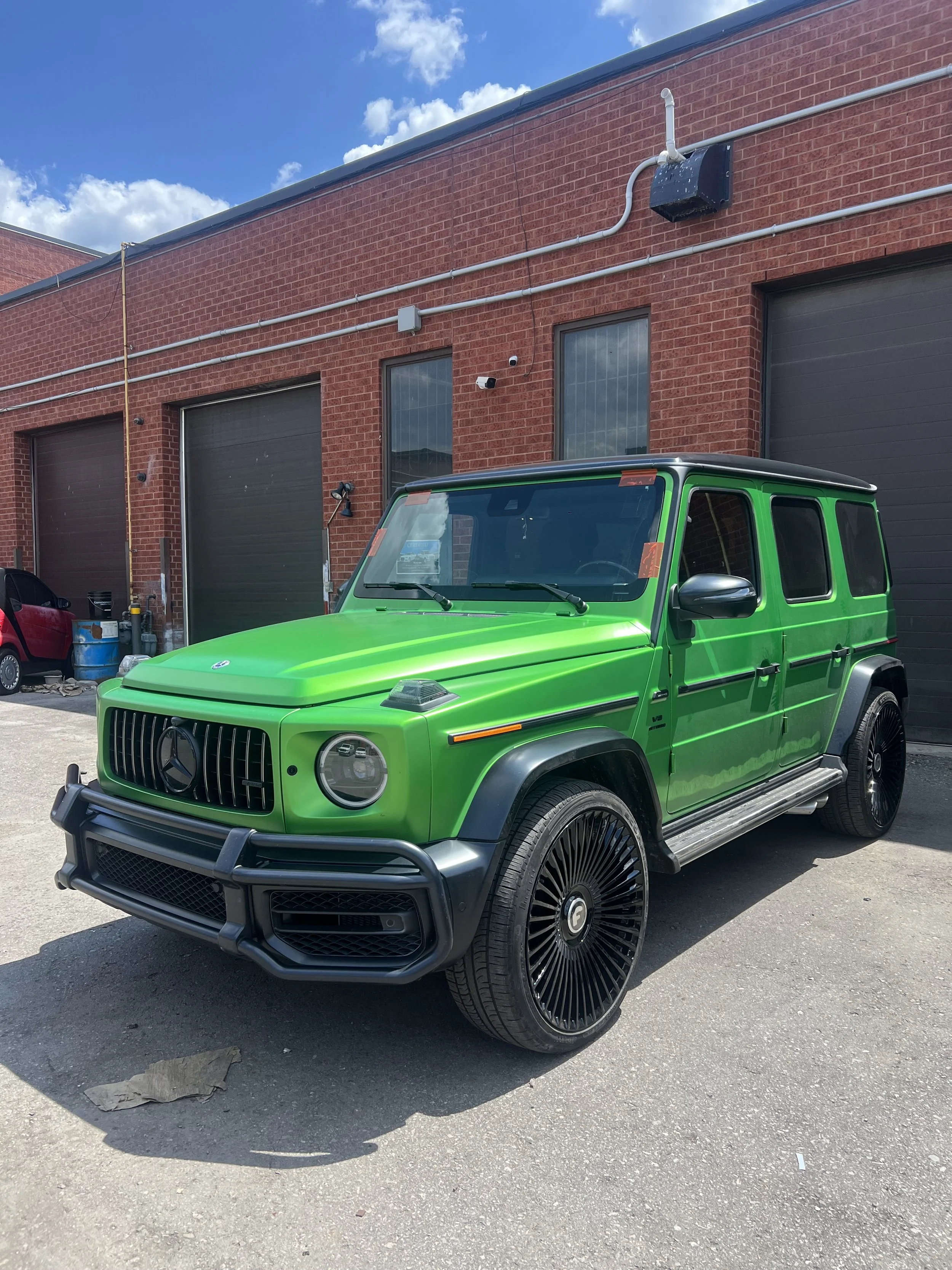 Green Mercedes-Benz G-Class SUV parked outside a red-brick building with black garage doors, under a partly cloudy sky.