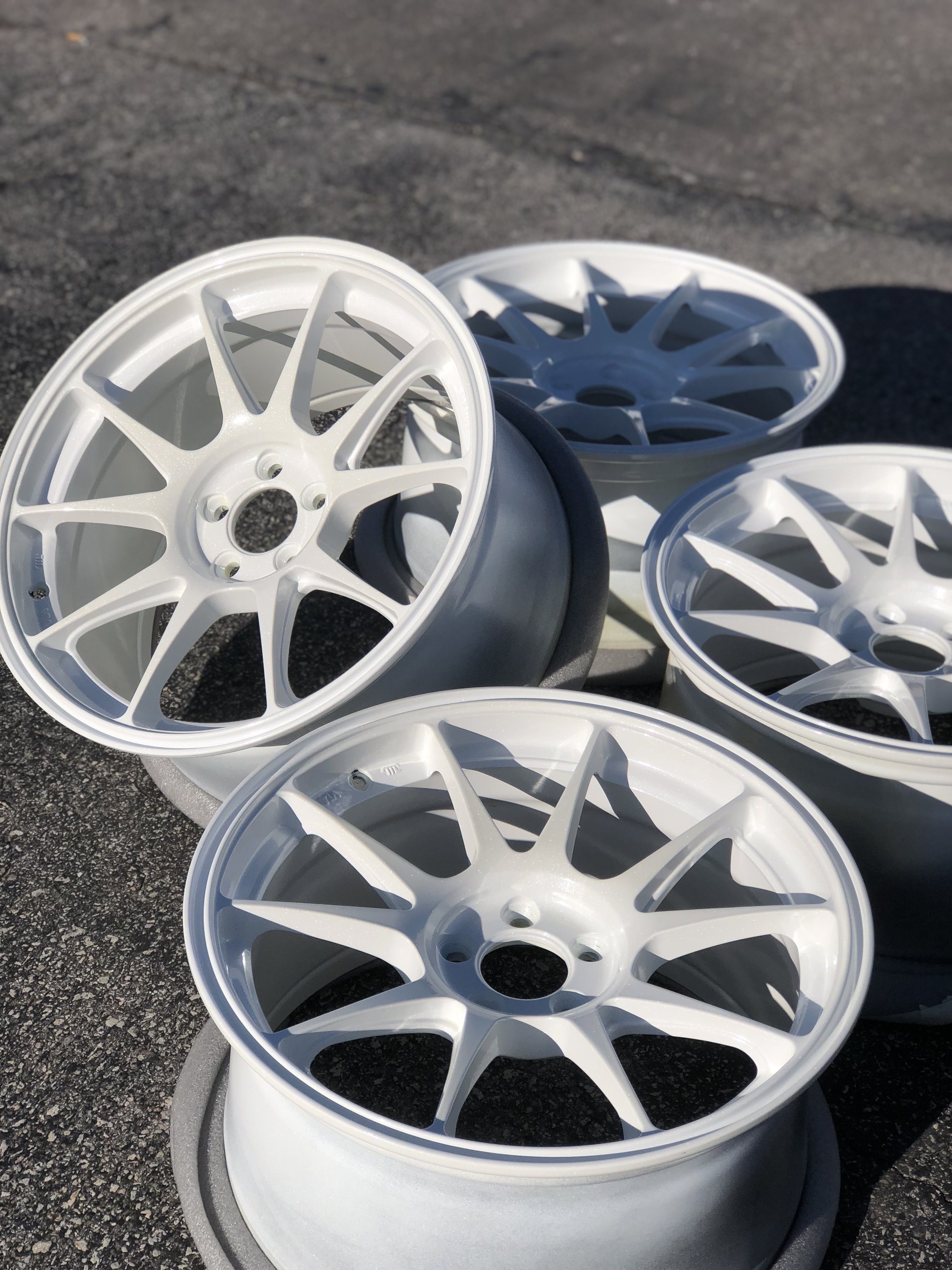 A set of five white car wheels with intricate spokes, placed on foam pads on an asphalt surface.