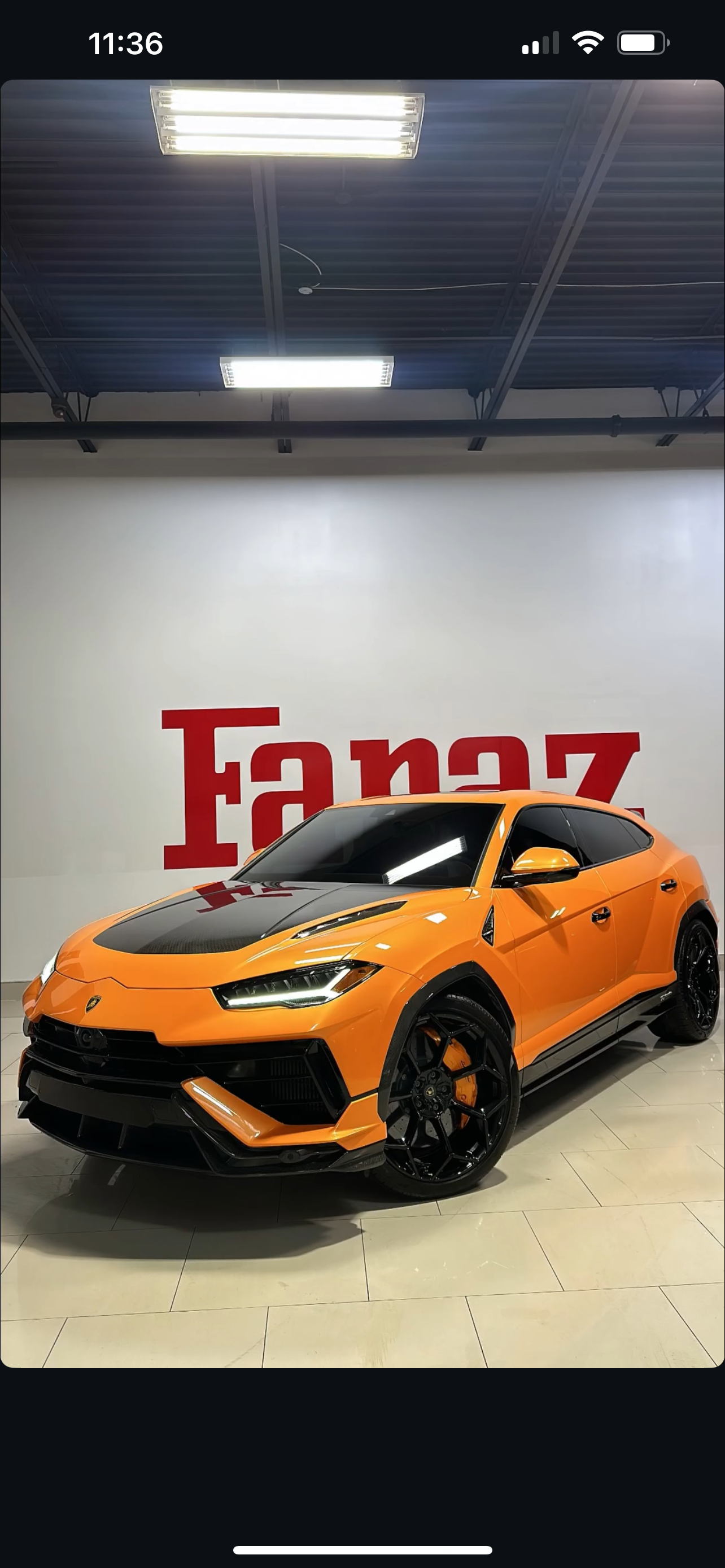 Orange Lamborghini Huracan with black accents inside a showroom, with a white wall displaying the letters 'Fanaz' in red behind it.