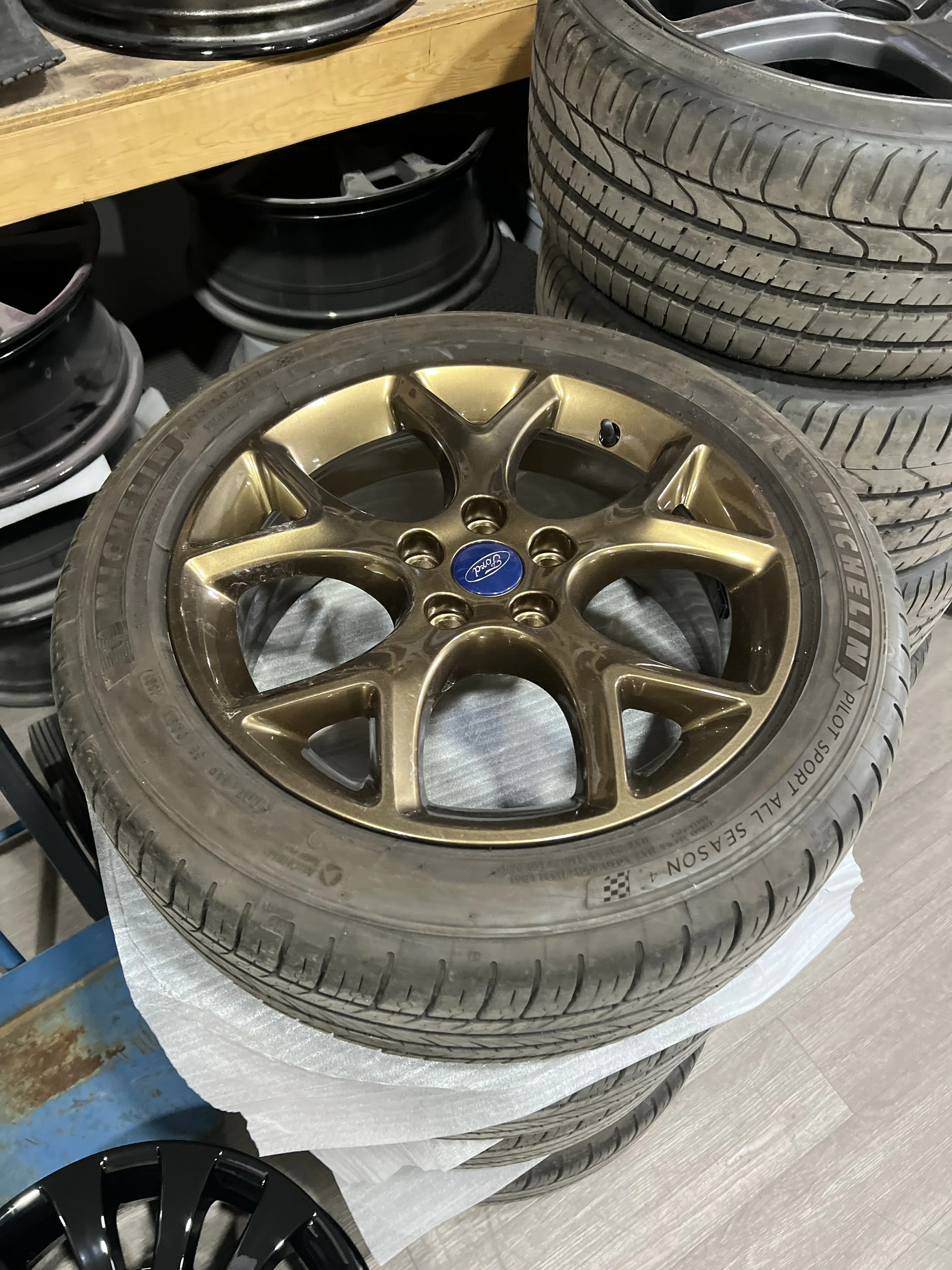 A set of four car tires with one mounted on a gold-colored alloy wheel with a blue Ford logo center cap, placed in a workshop with other wheels and tires in the background.