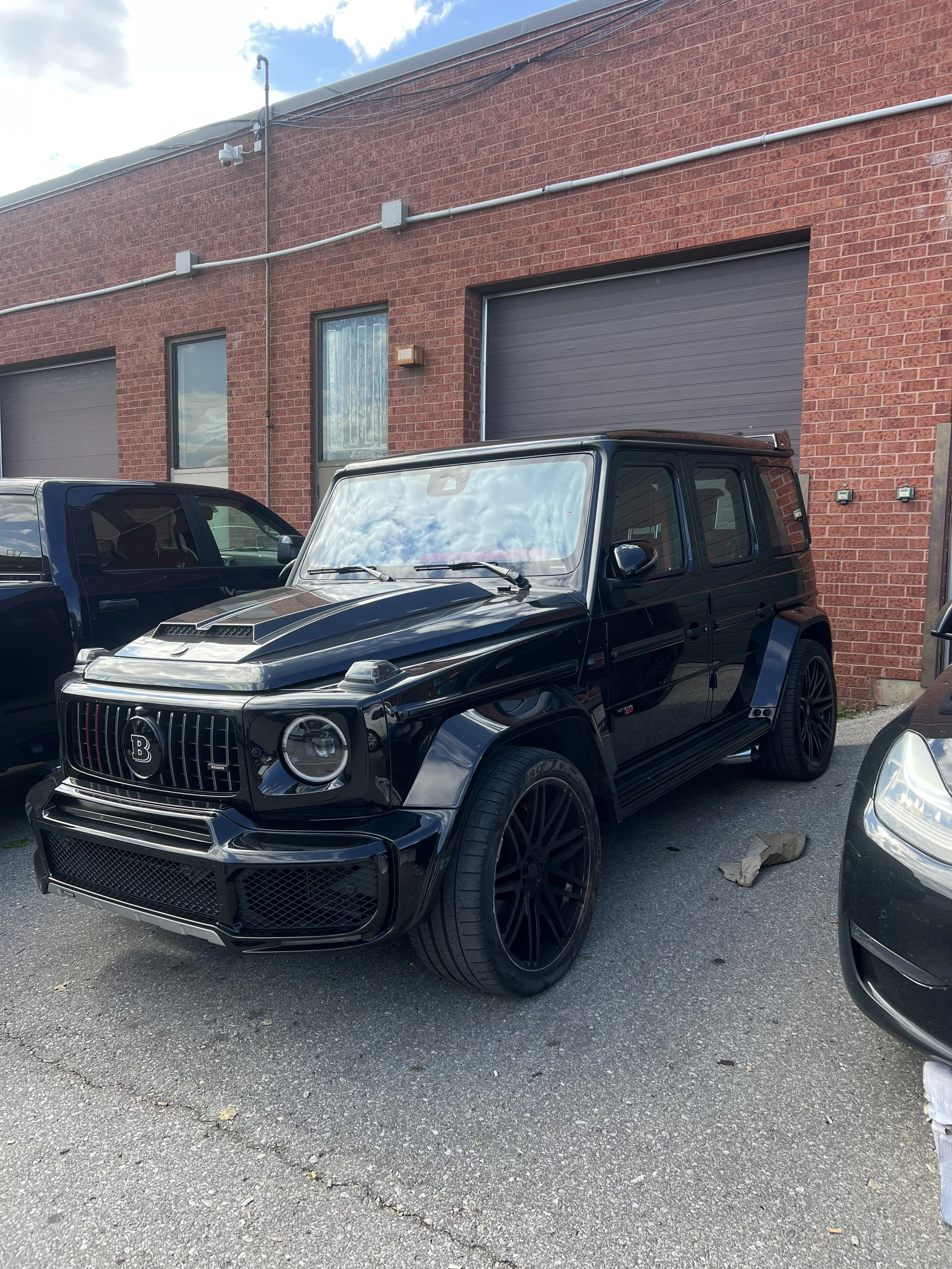 A black modified luxury SUV with a main grille featuring a 'B' emblem, large mesh grille and blacked out wheels, parked in front of a red brick building with closed garage doors.