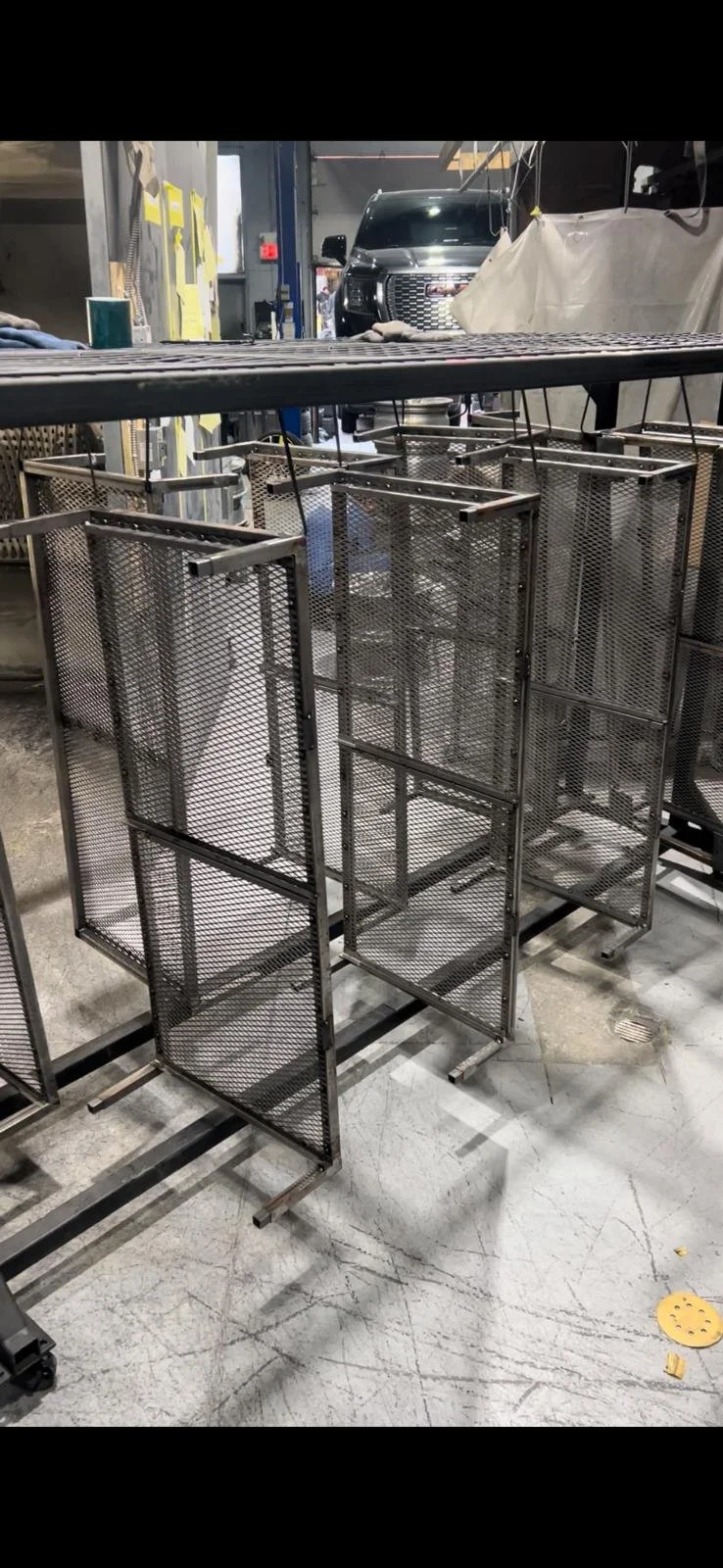 Empty metal mesh display racks on wheels inside an industrial workspace or store, with a car visible in the background.