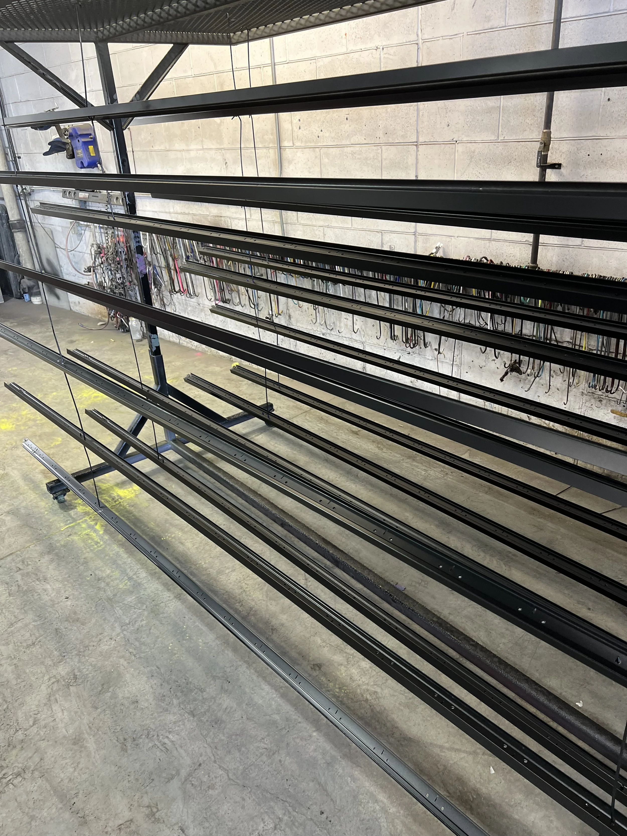 Image shows a metal shelving unit with empty track rails for a sliding or rolling mechanism, located in an industrial or workshop setting with tools hanging on the wall.