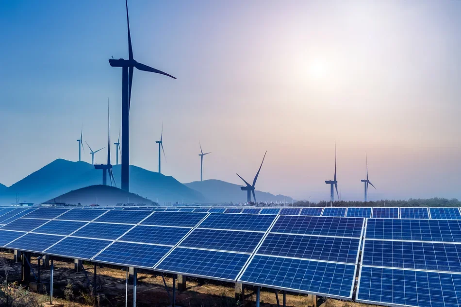 Solar panels in the foreground and wind turbines on hills in the background under a hazy sky.