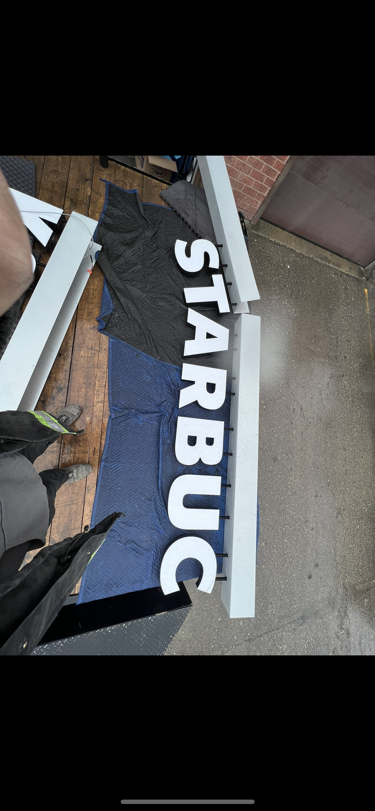 The image shows a sign that reads 'START BUILD' lying on a workspace with tools and materials for assembly, with a person standing nearby.