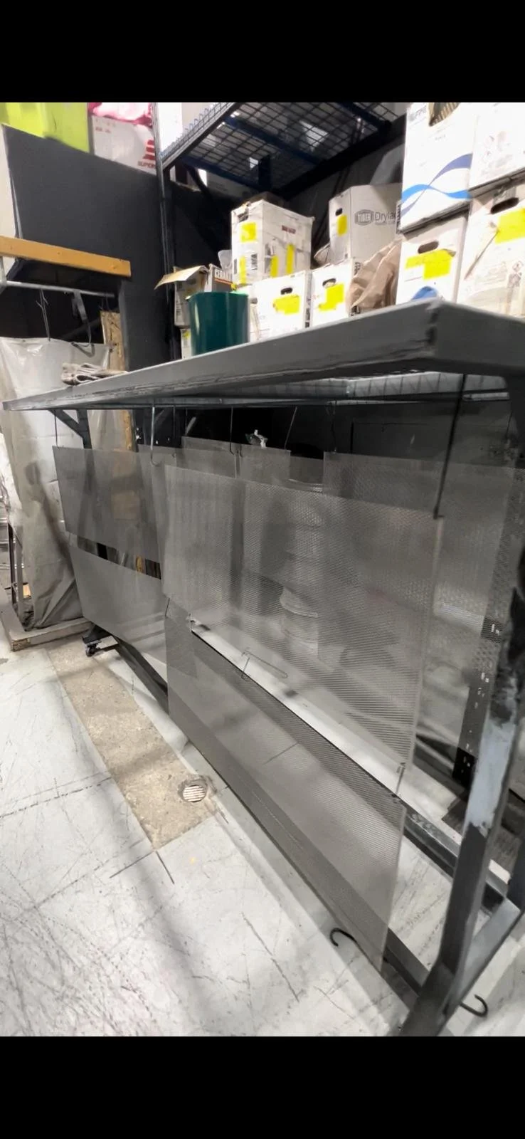 A stainless steel commercial oven or warming drawer unit on wheels inside a storage or warehouse space with boxes and items on a high shelf in the background.