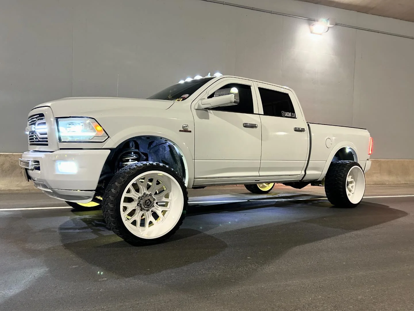 A white pickup truck with large custom white wheels parked in a tunnel with gray walls. The truck features bright headlights, LED lights on the roof, and tinted windows.