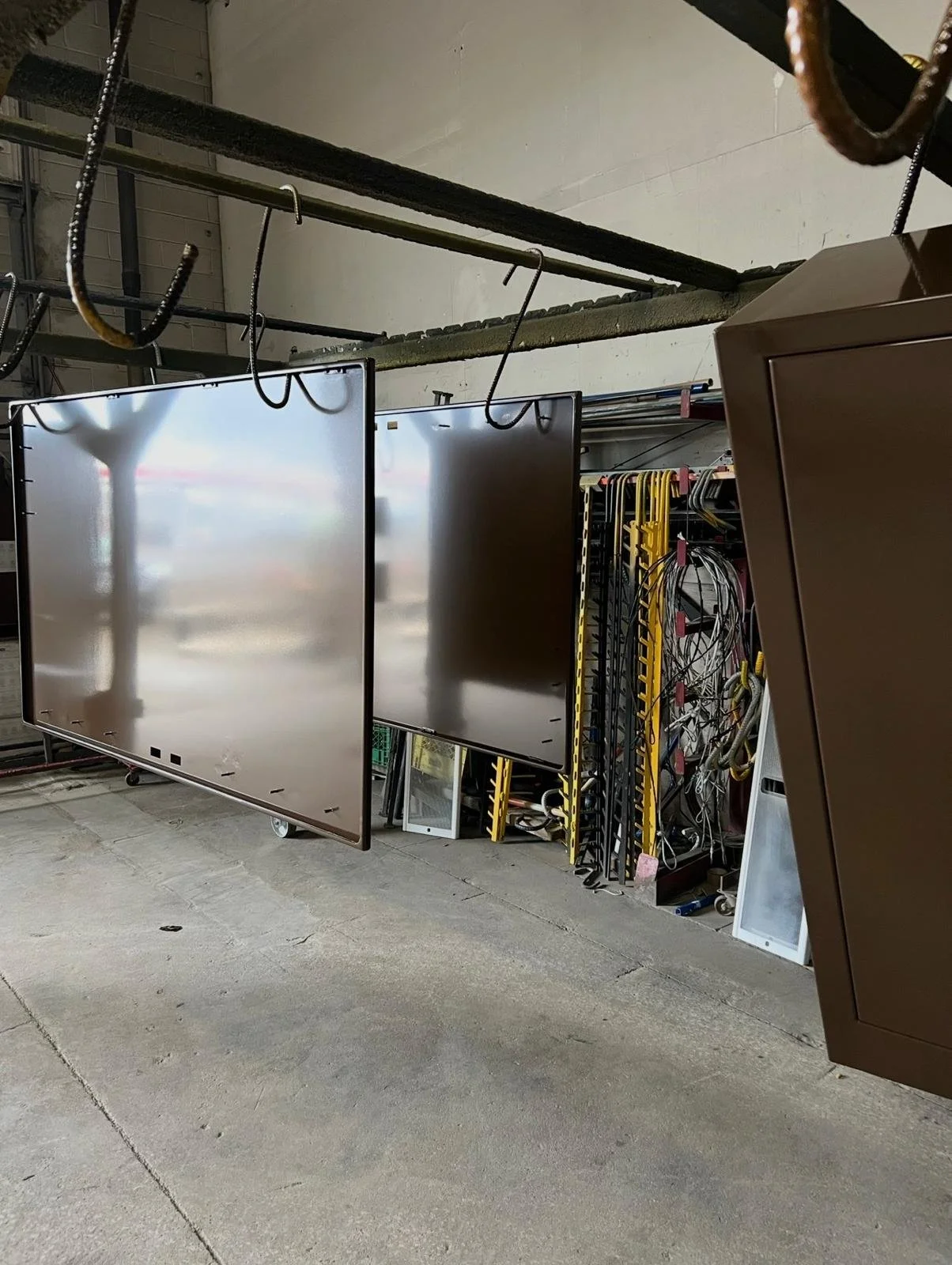 Multiple large flat screen monitors hanging from ceiling hooks in a warehouse or storage room, with some electronic equipment and cables visible behind.