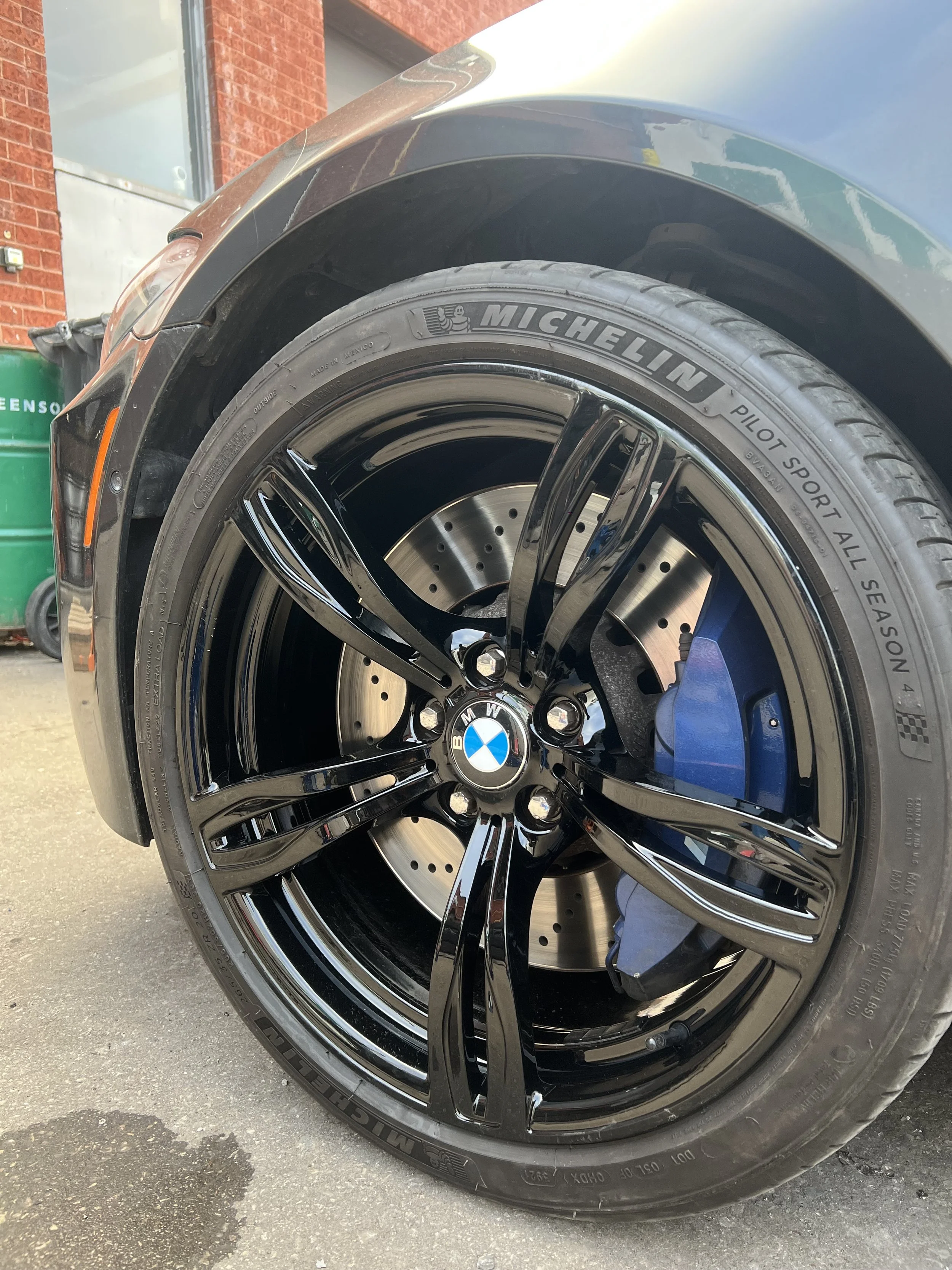 Close-up of a black BMW car wheel with Michelin Pilot Sport All Season tire, showing a black alloy rim with the BMW logo in the center, blue brake caliper behind the rim, and a drilled brake disc.