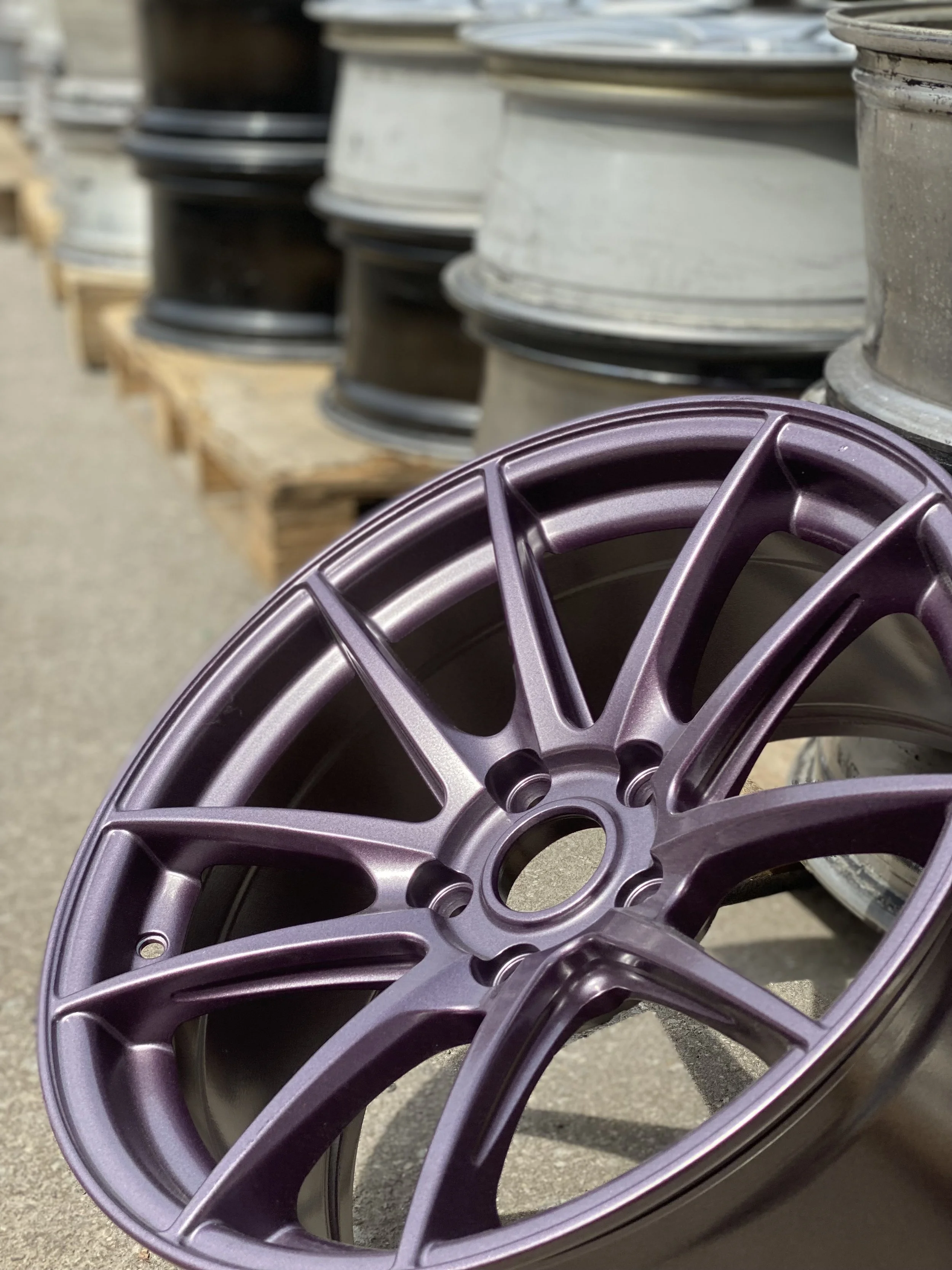 Close-up of a purple alloy car wheel rim with other wheels stacked in the background at an outdoor location.