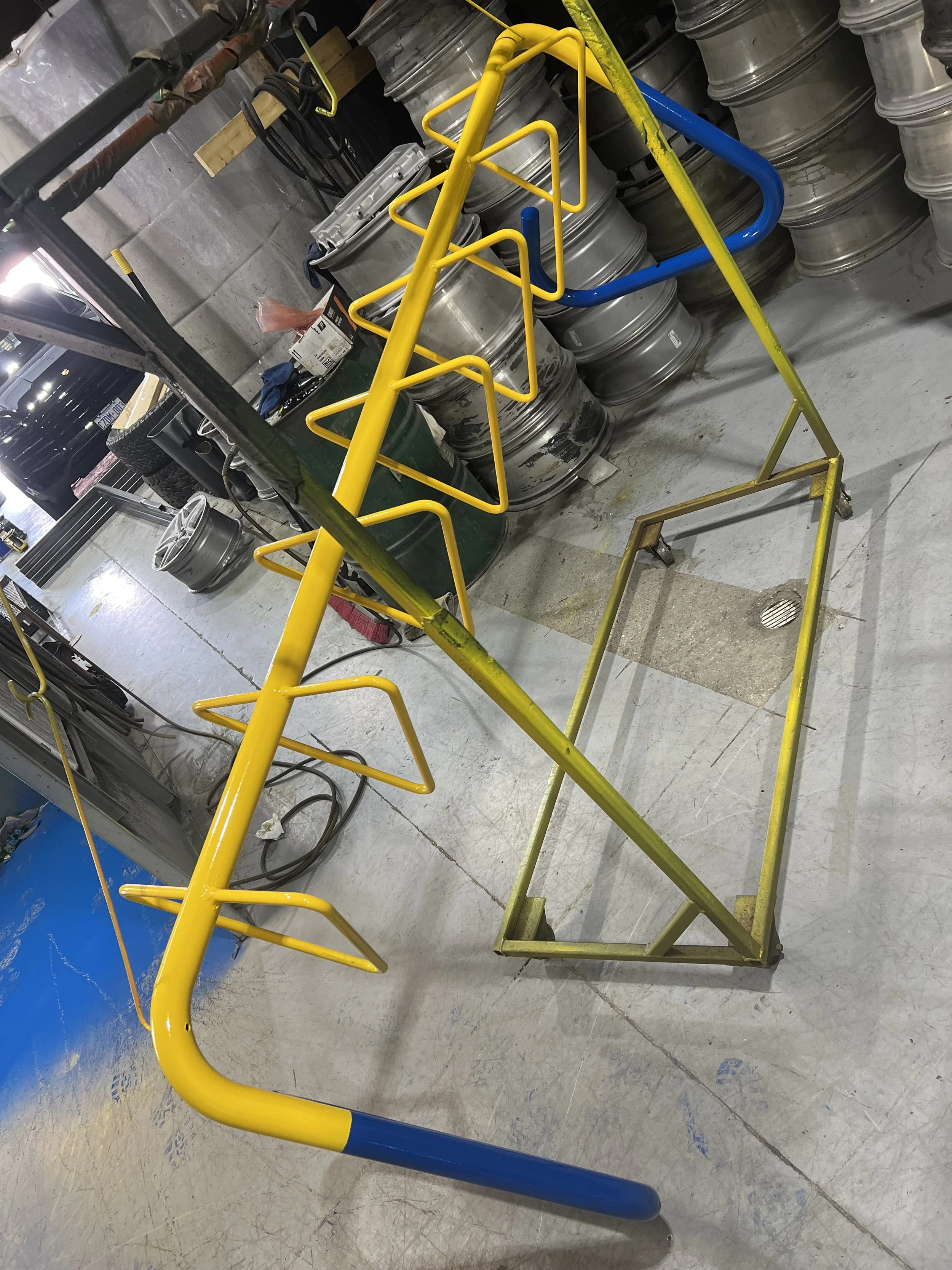 Metal yellow and blue cart with hooks, placed in a workshop or garage environment with metal wheels and various tools and equipment in background.