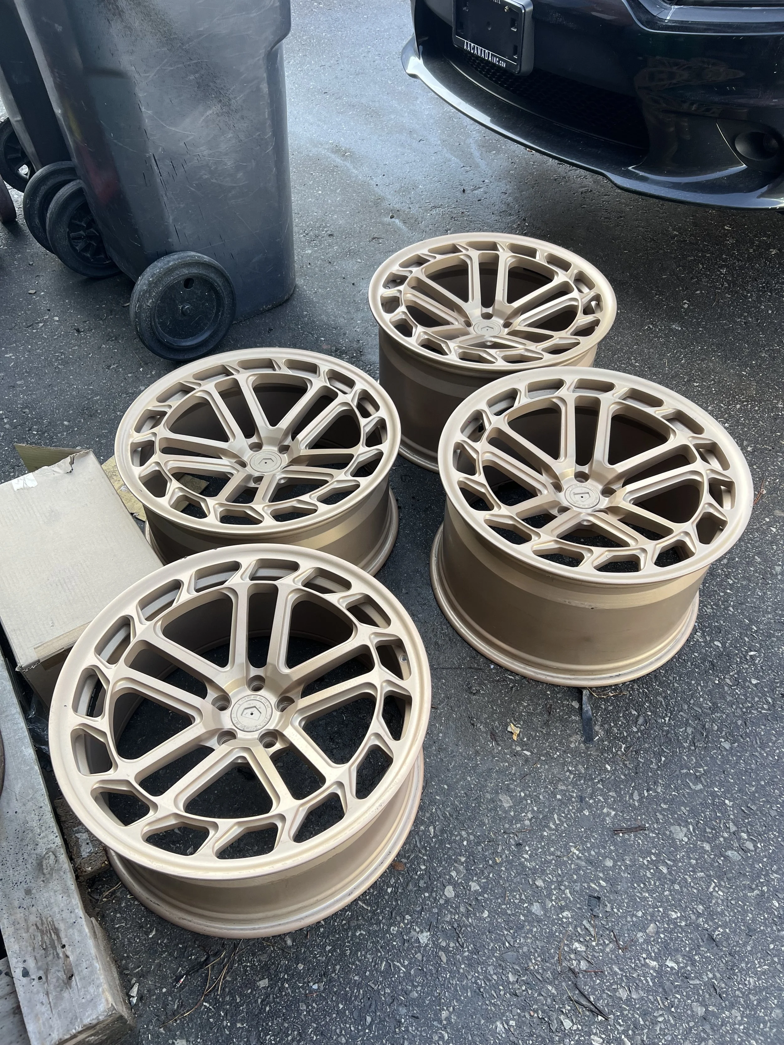 Four beige-colored car wheels stacked outdoors on asphalt near a black vehicle and a trash bin.