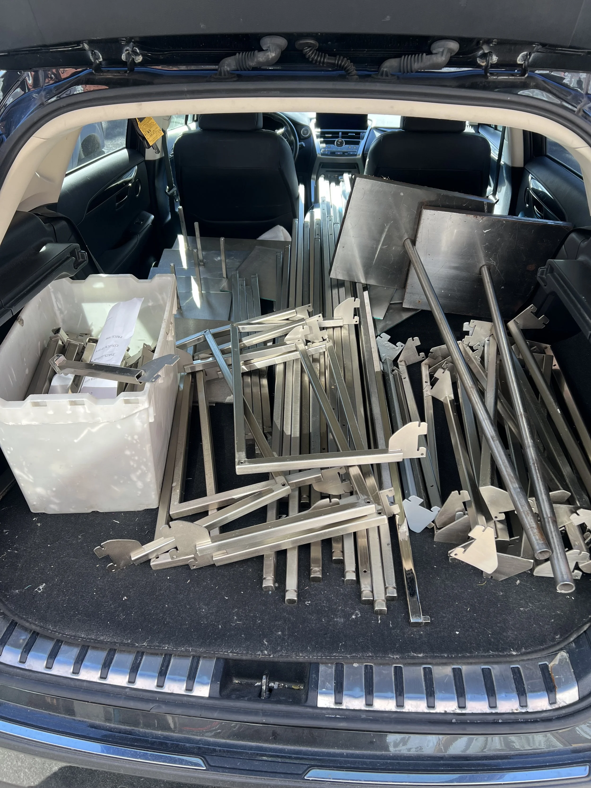 The trunk of a car filled with metal parts, tools, and a plastic container, suggesting assembly or repair work.