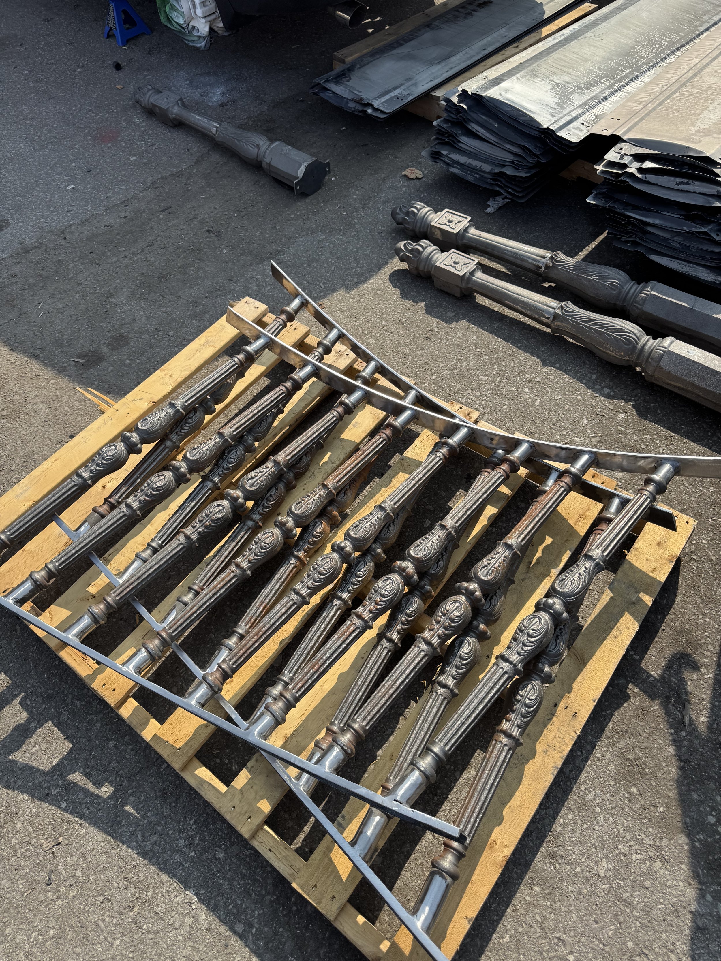 A pallet of ornate metal fence posts and rails, with additional fence parts stacked nearby, arranged outdoors on an asphalt surface.