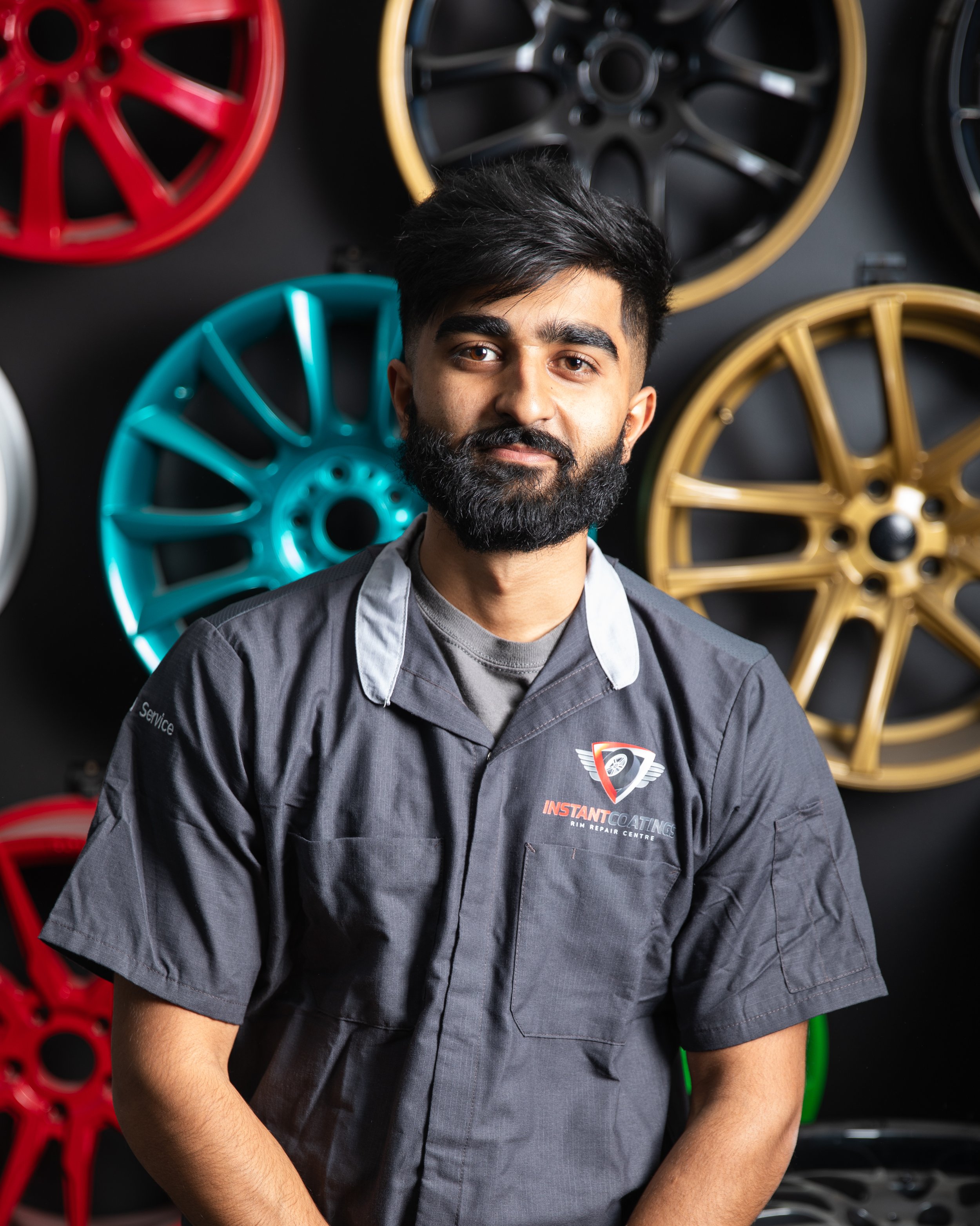 A man with a beard and dark hair smiling at the camera, wearing a gray mechanic's uniform with a logo, standing in front of a wall with colorful alloy wheels.