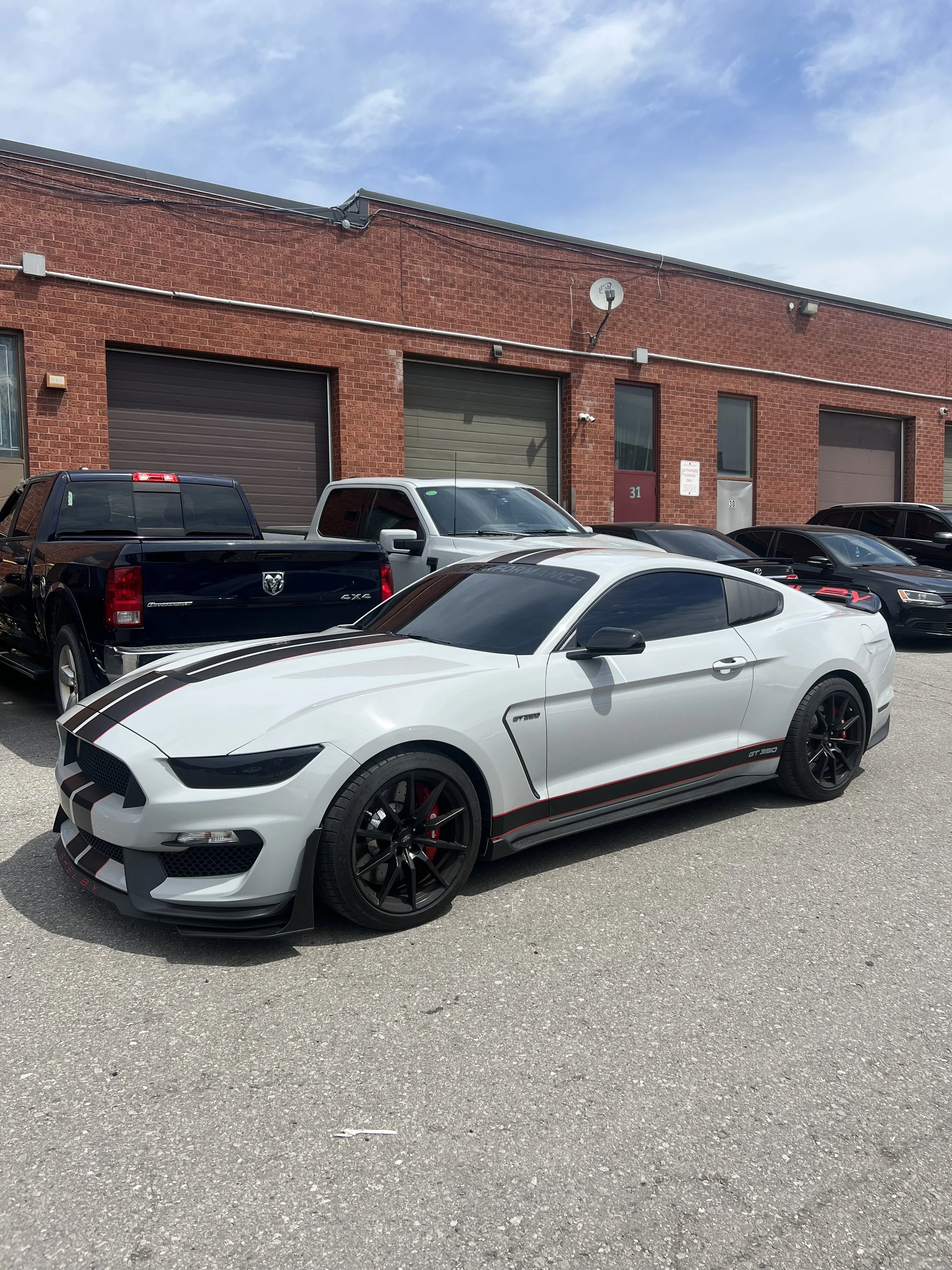 A white sports coupe with black stripes and red accents parked in front of a brick building, surrounded by other vehicles.