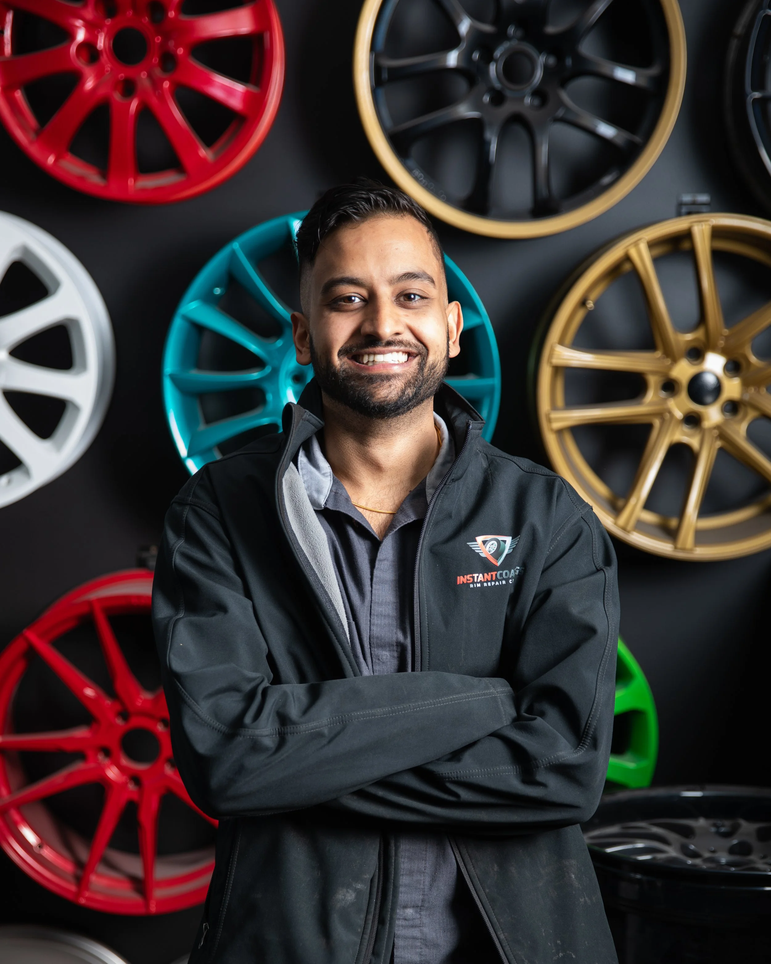 A smiling man with crossed arms standing in front of colorful car rims mounted on a wall.