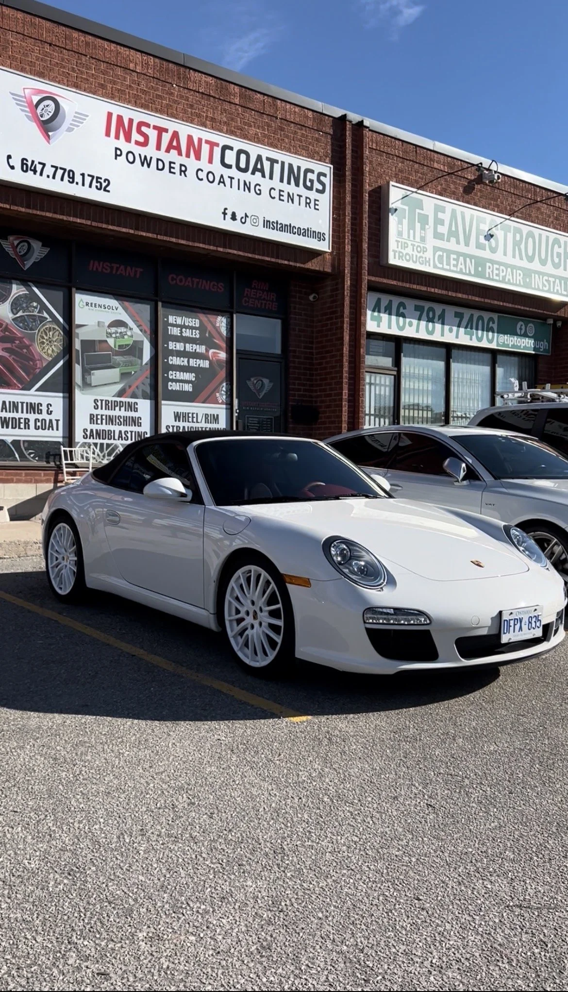 White Porsche sports car parked in front of a building with signs for a powder coating center and an auto repair shop.