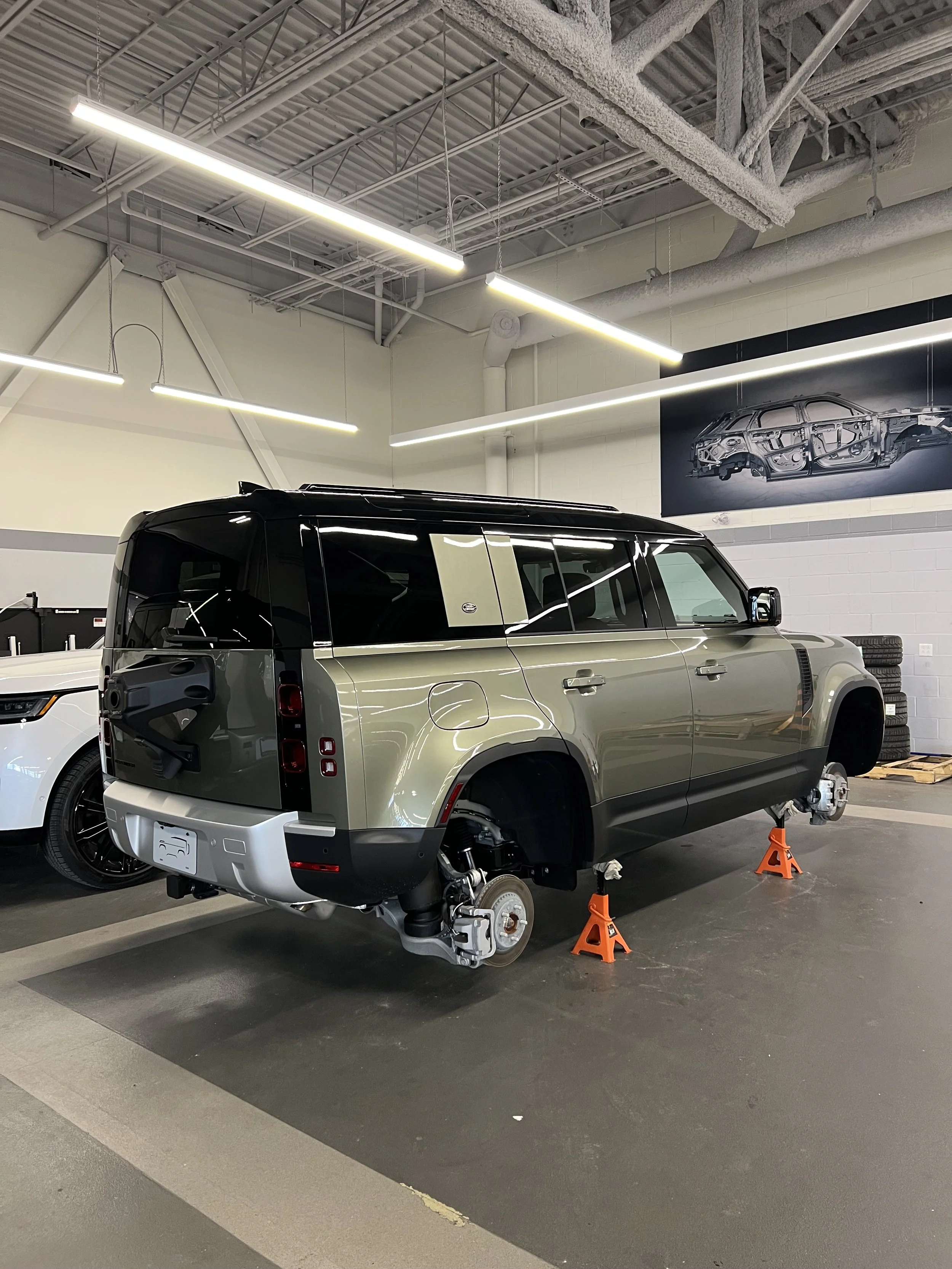 A Land Rover vehicle is elevated on orange jack stands inside a car workshop, missing its wheels, with a white car parked nearby. There is a large poster of a car manufacturing diagram on the wall.