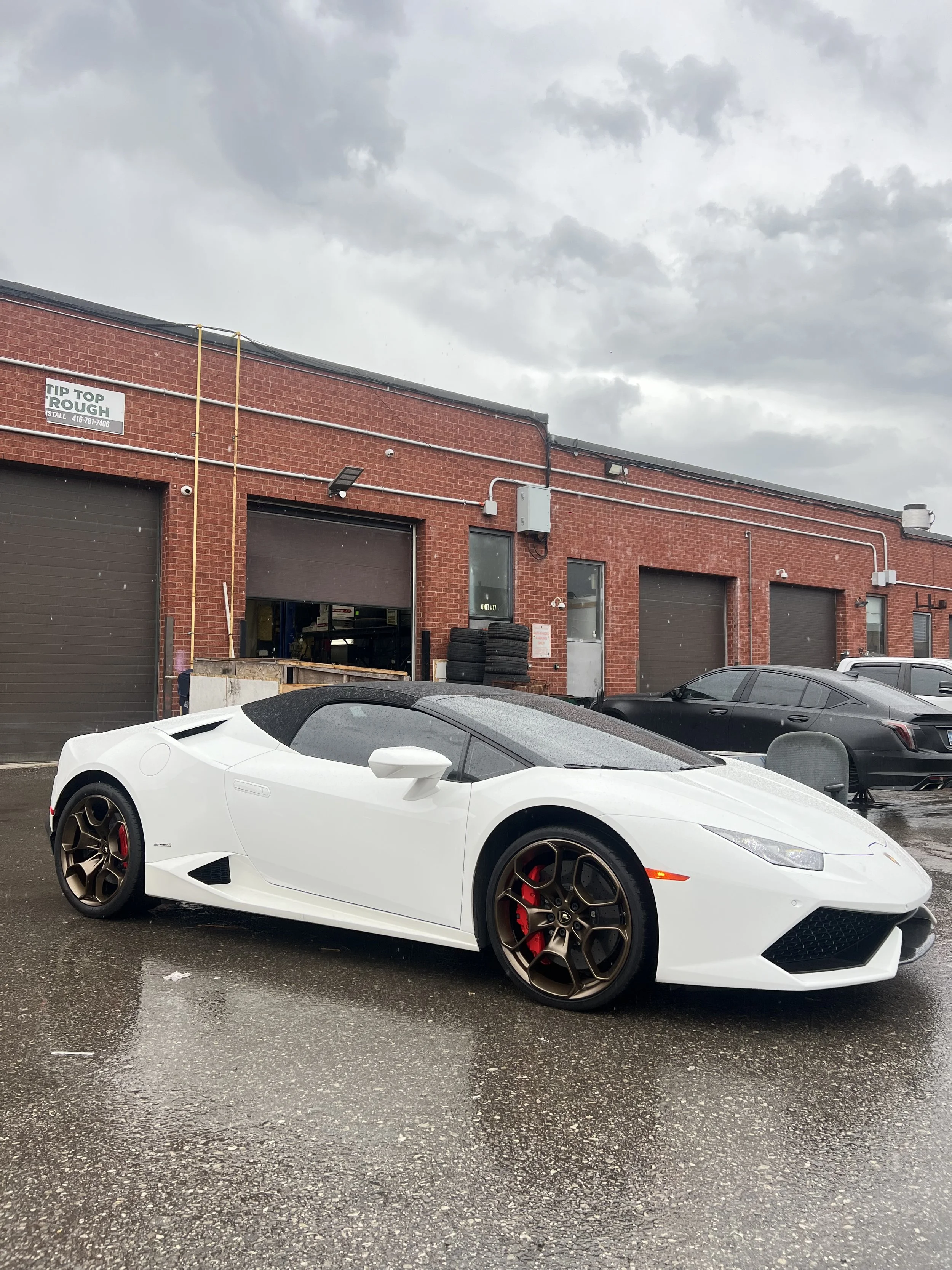 White convertible sports car parked outside in front of a brick building on a rainy day.