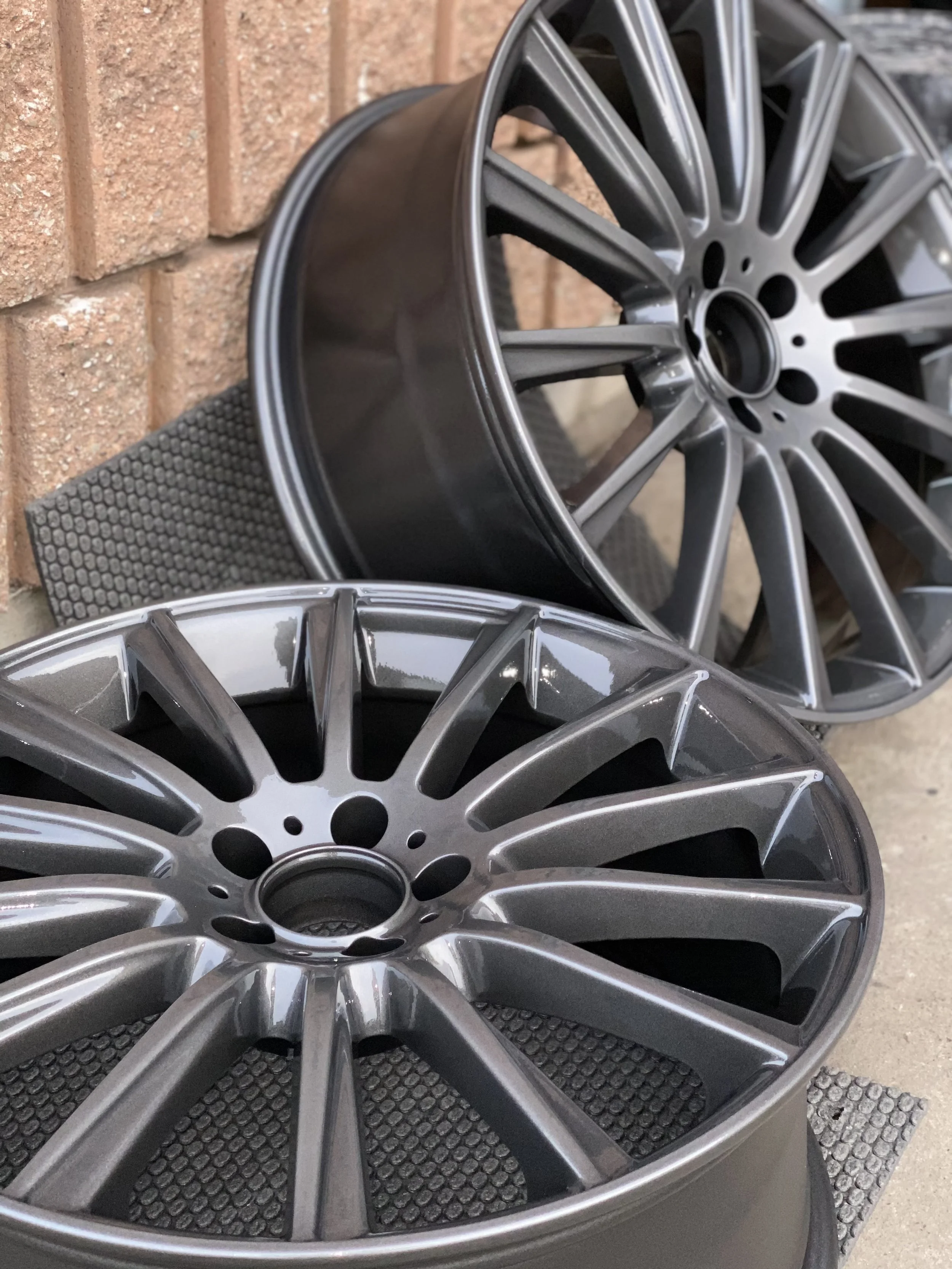 Two new metallic gray car wheels with intricate spoke design resting on a black rubber mat against a brick wall.