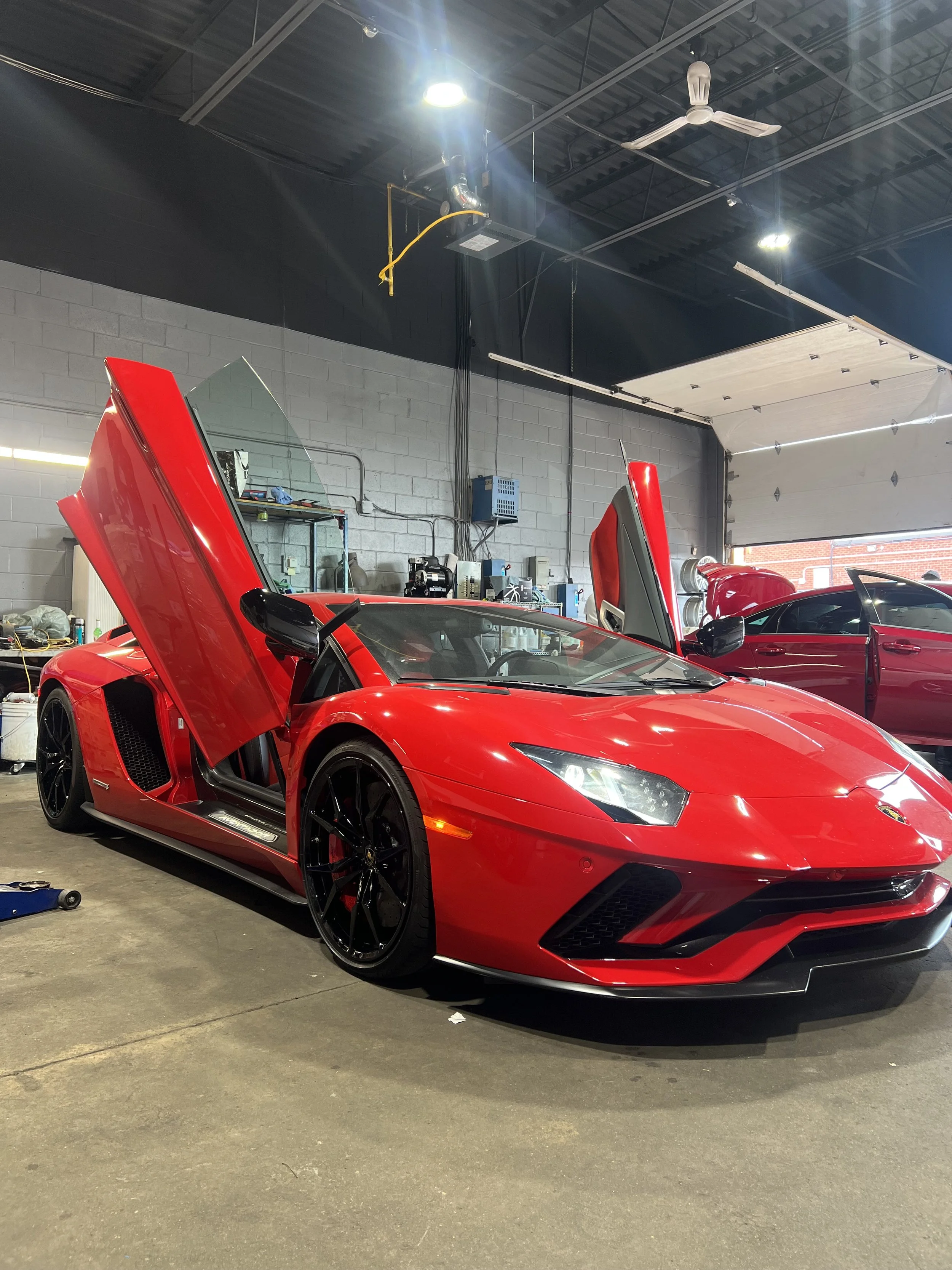 Red Lamborghini sports car with scissor doors open inside a garage or workshop with various equipment and tools visible in the background.