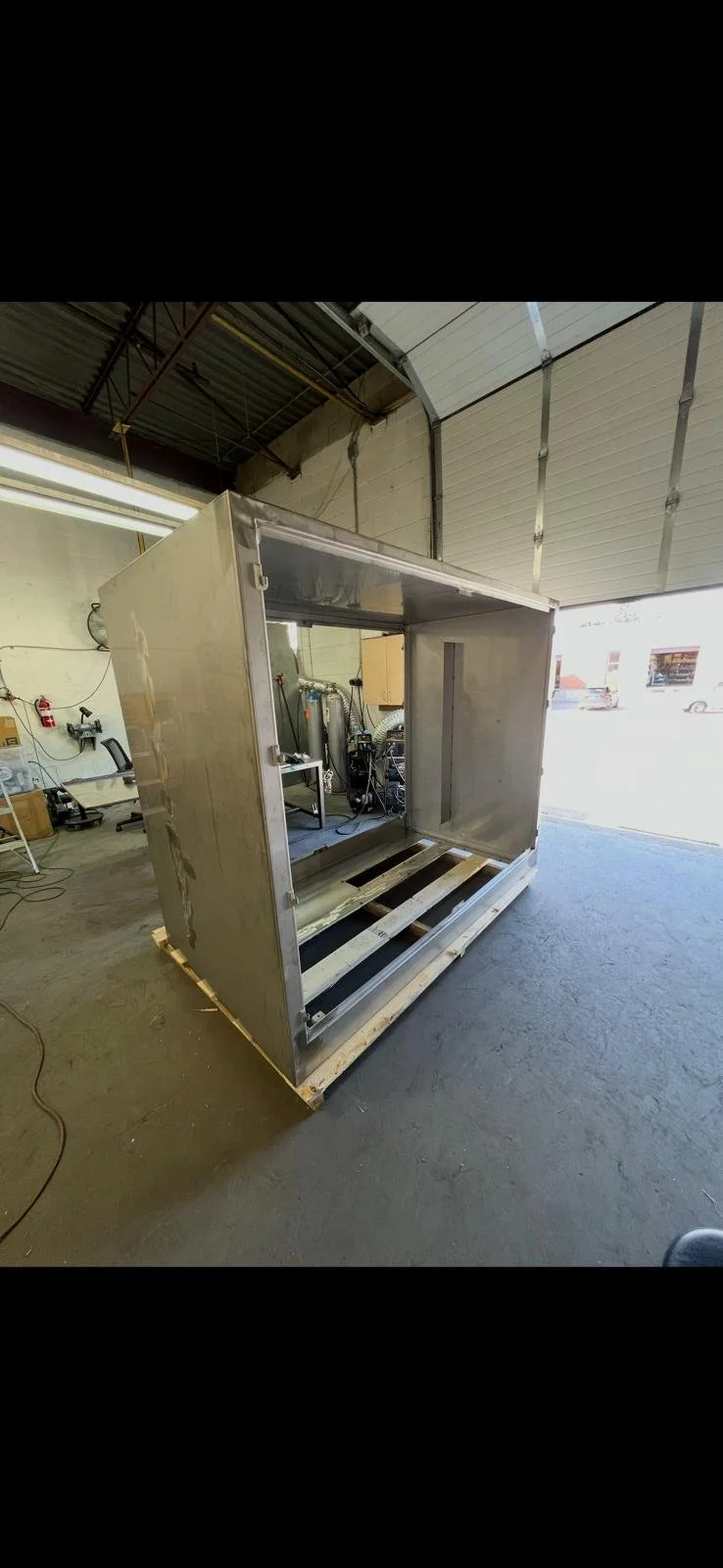 Unfinished metal enclosure or frame inside a warehouse, with wooden pallets at the base and tools and equipment in the background.