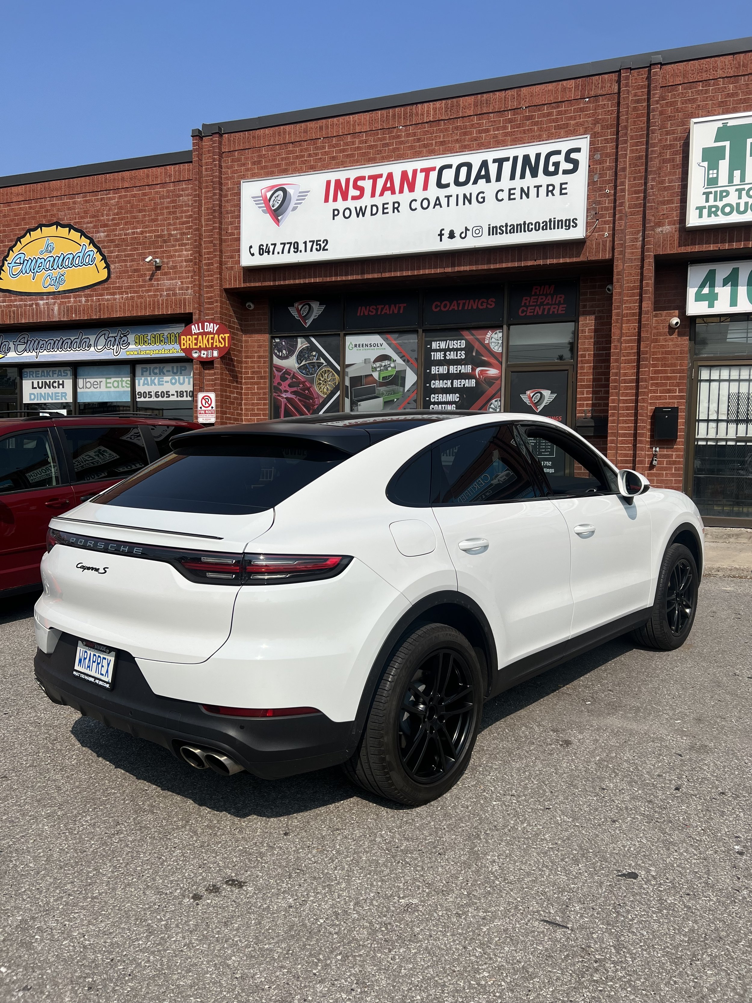 A white Porsche Macan S SUV parked in front of a storefront with various signage, including 'Instant Coatings' for powder coating and rim repair center, and 'La Empanada Cafe' with a sign for breakfast all day.