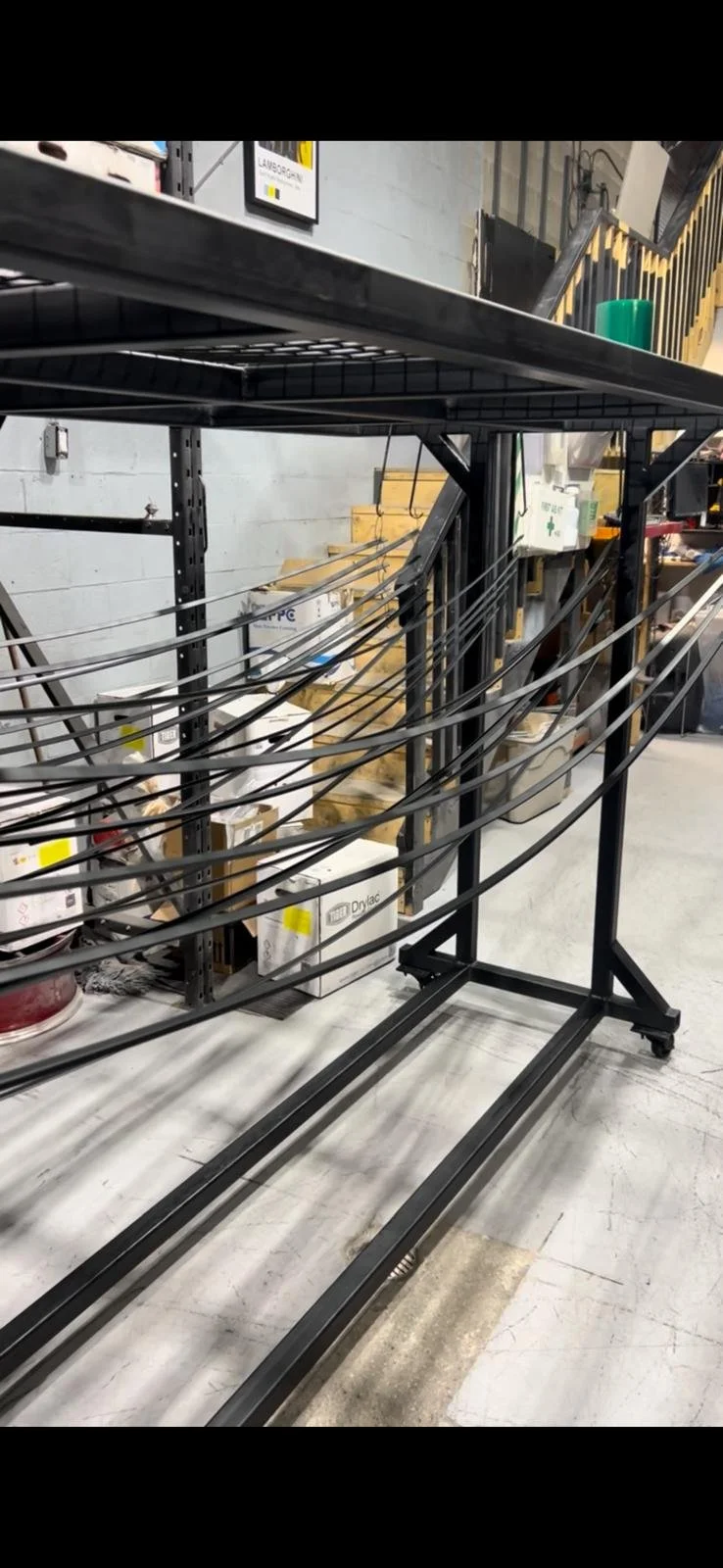 Empty black metal retail or storage cart with horizontal wires, on a warehouse or store floor with shelves and supplies in the background.