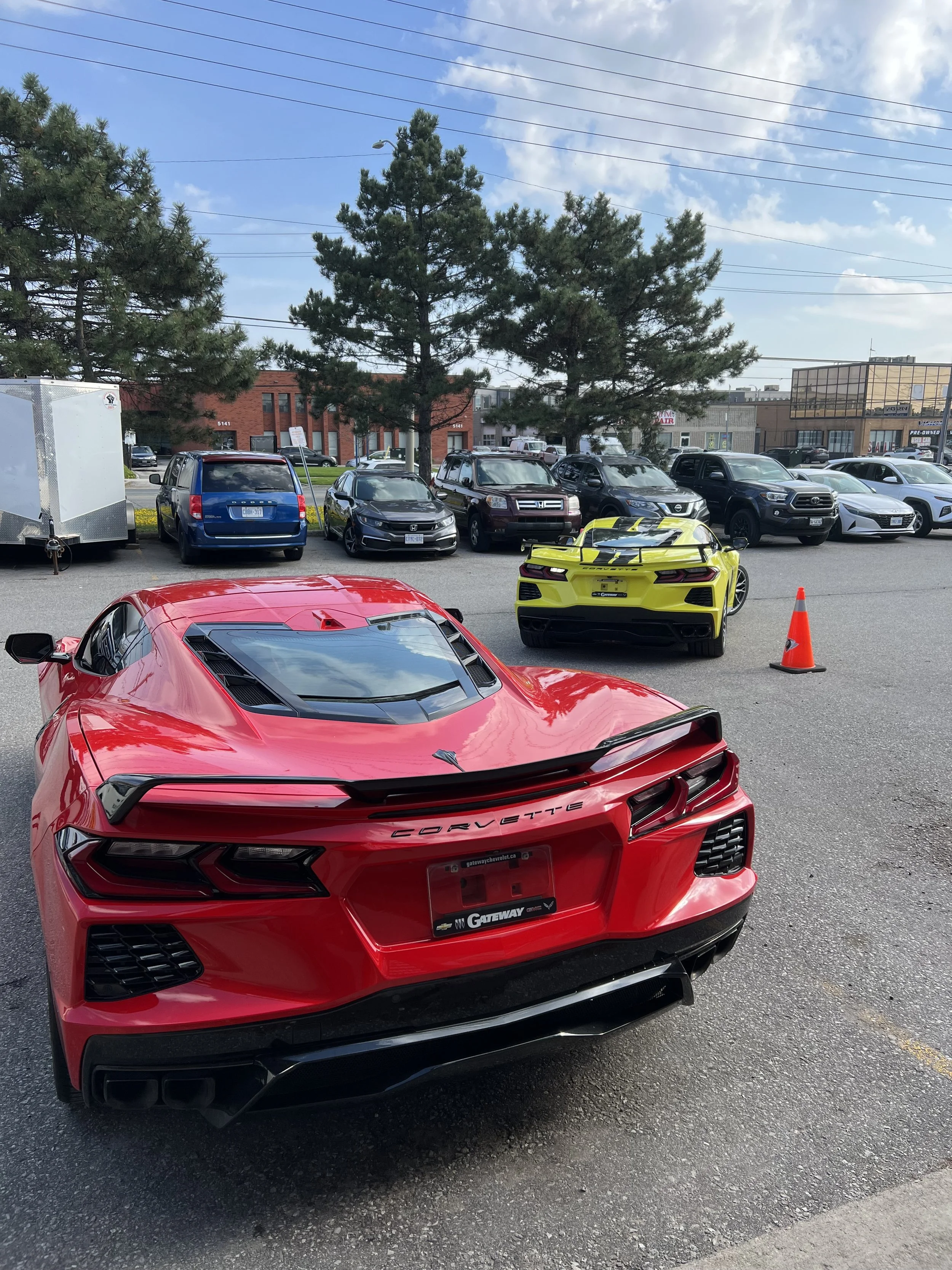 A red Chevrolet Corvette sports car parked in a lot with a yellow sports car in front and several cars parked in the background.