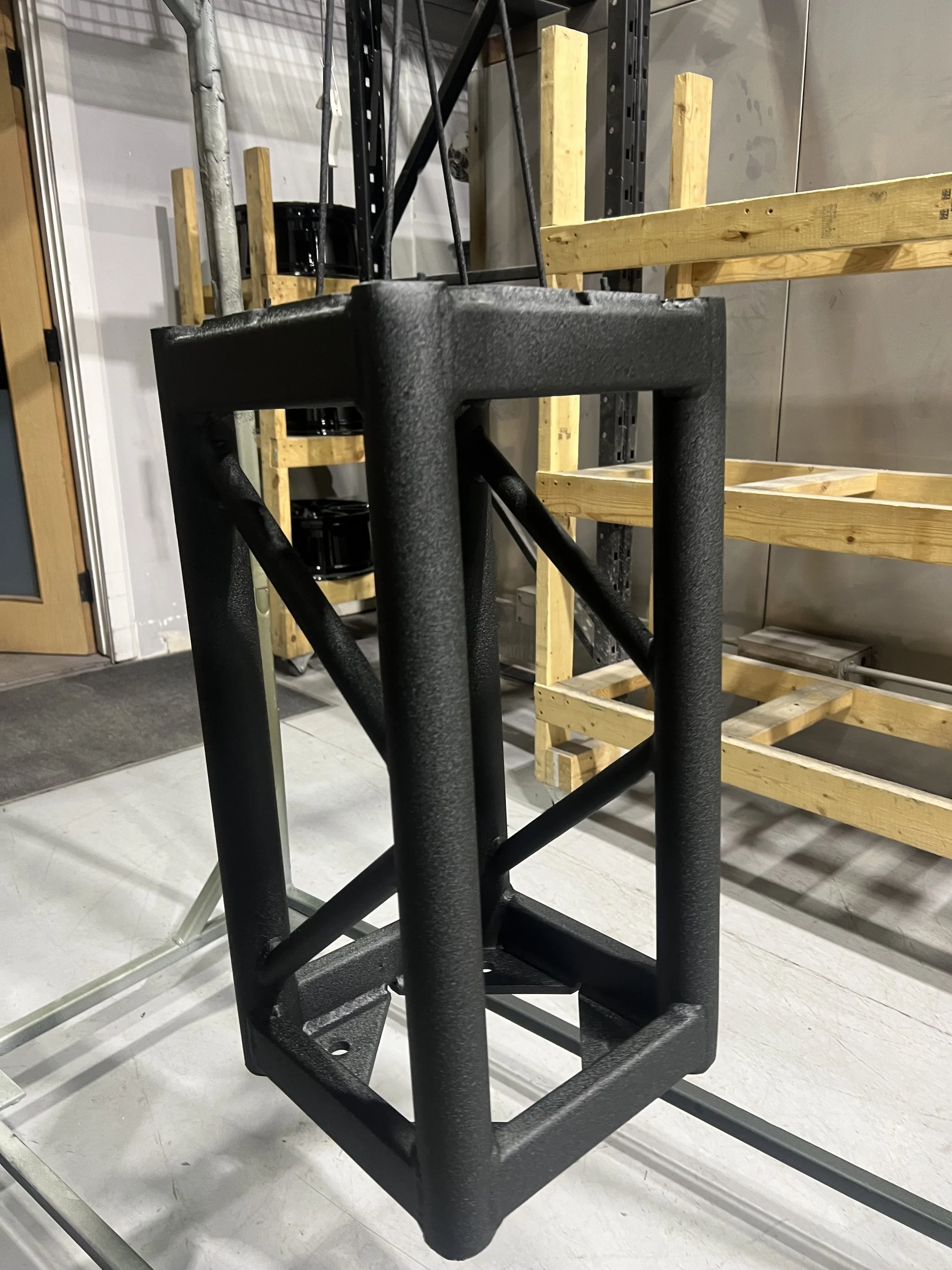 A black metal frame structure with angled supports, standing on the floor in a storage room with shelves and wooden pallets.