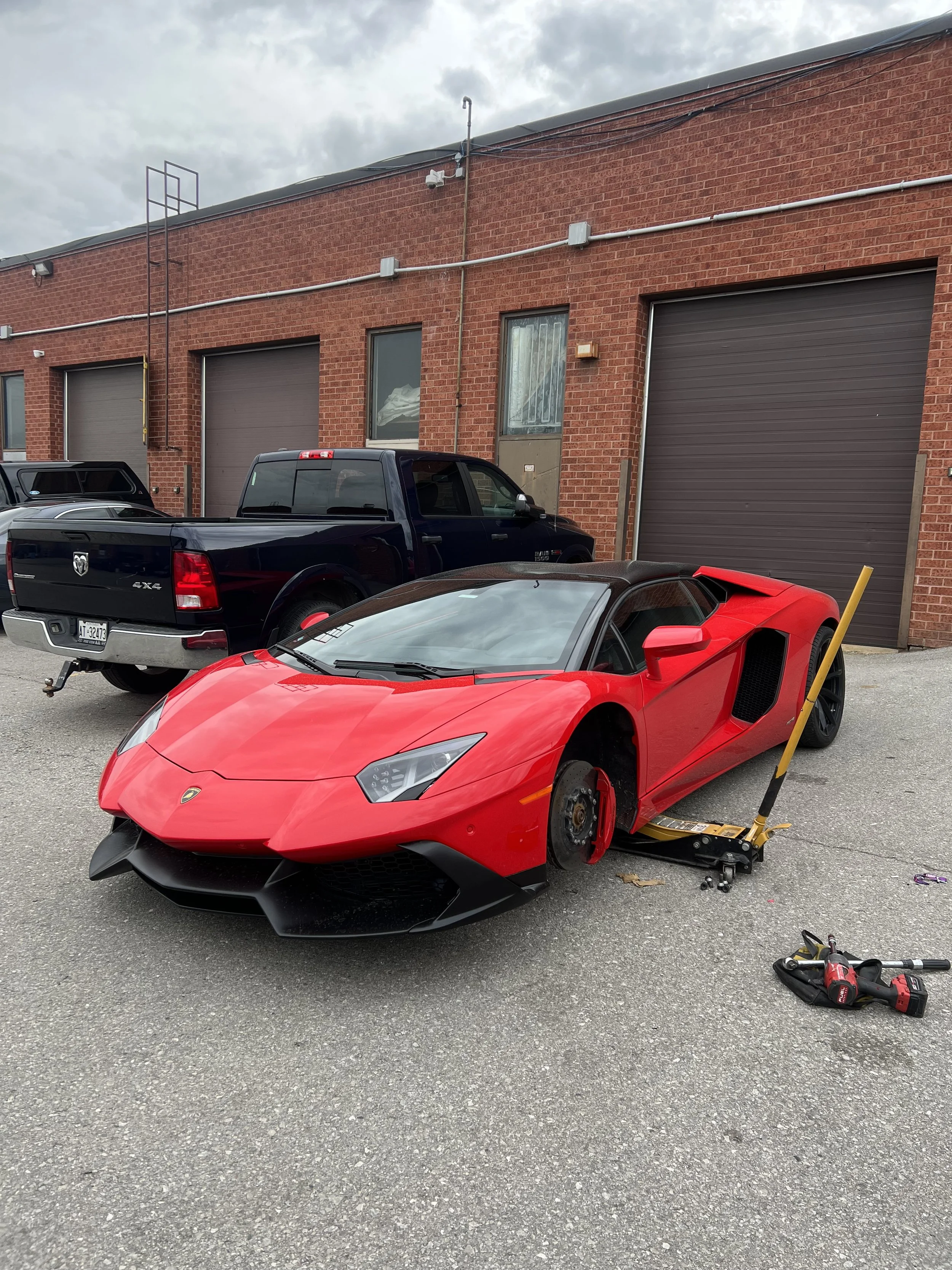 Red Lamborghini sports car with front wheel removed, jack and tools nearby, parked outside brick building with garage doors, gray cloudy sky.