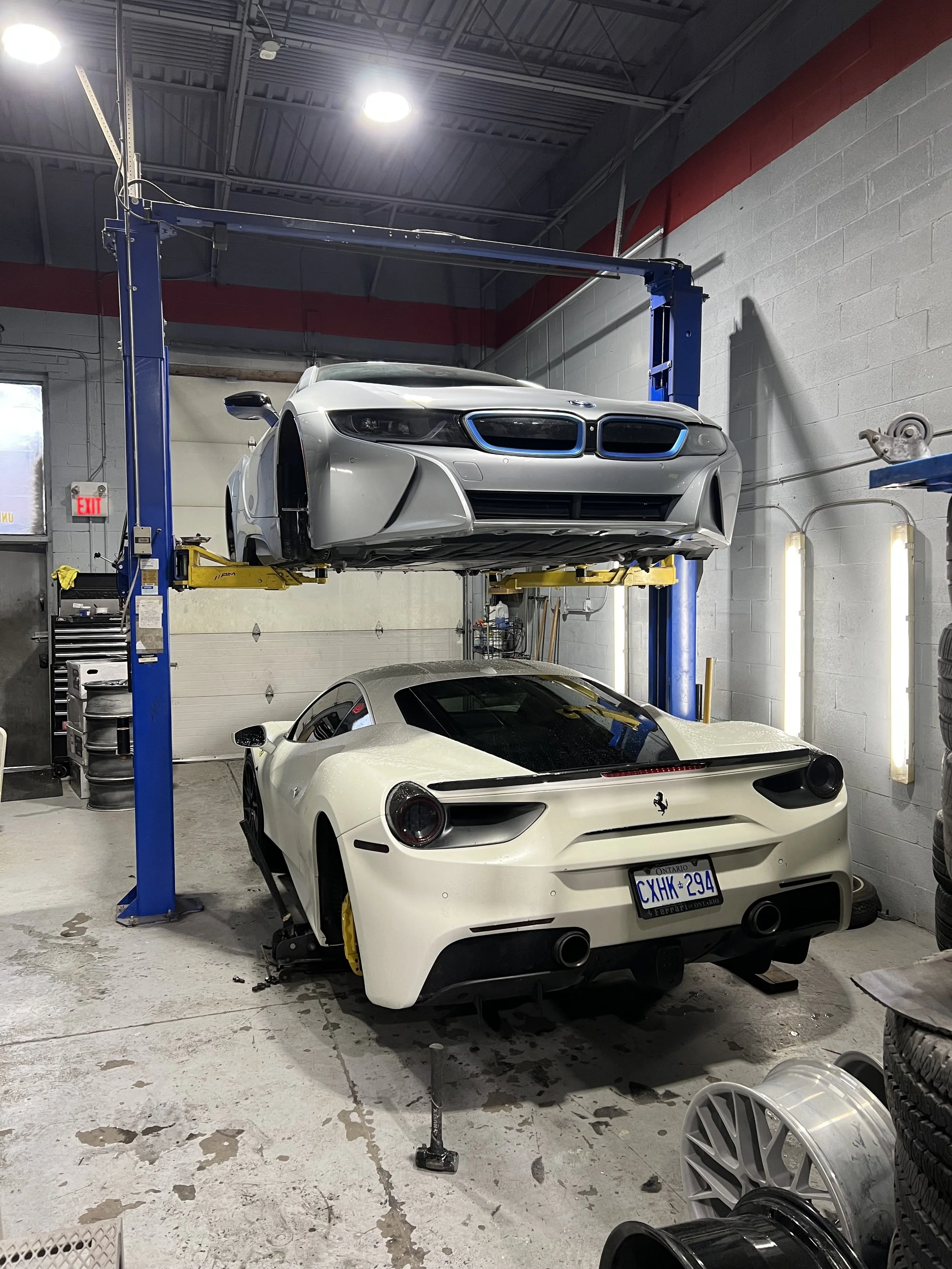 Two sports cars in a garage, one white and one silver, with the silver car lifted on a hydraulic lift.