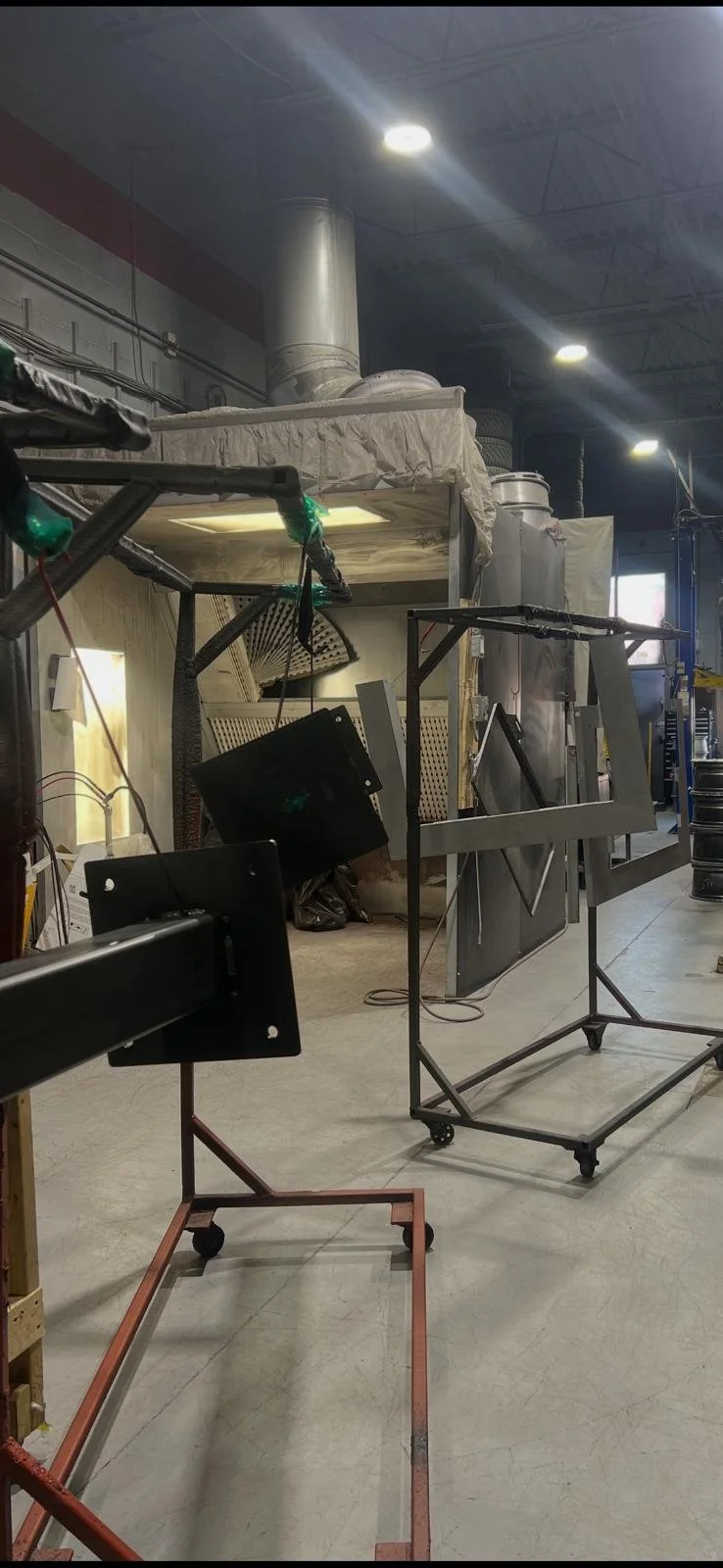 Industrial workshop with metal frames, machinery, and ventilation pipes, illuminated by ceiling lights.