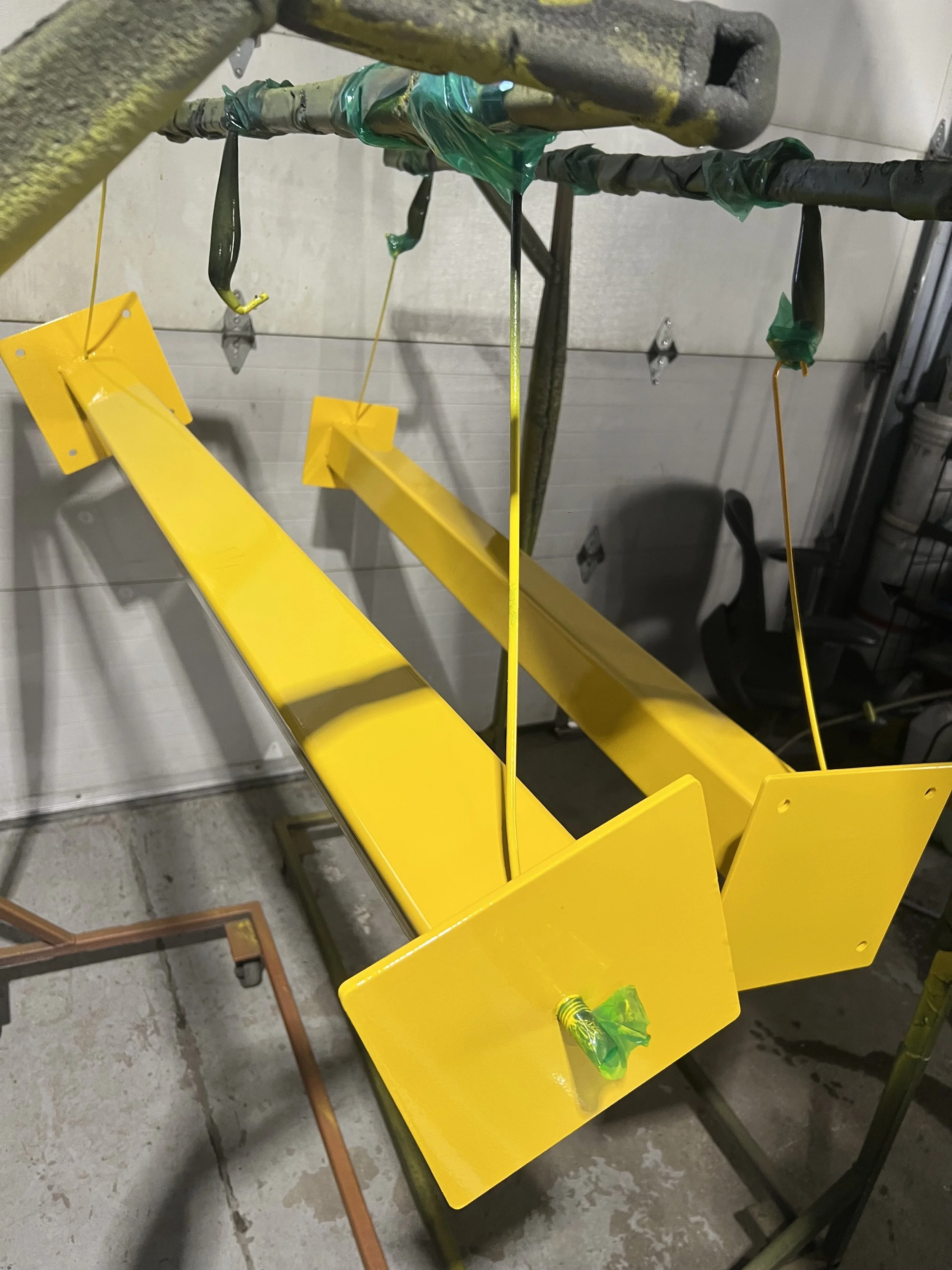 A yellow-painted metal frame being painted, hanging from hooks in a workshop.