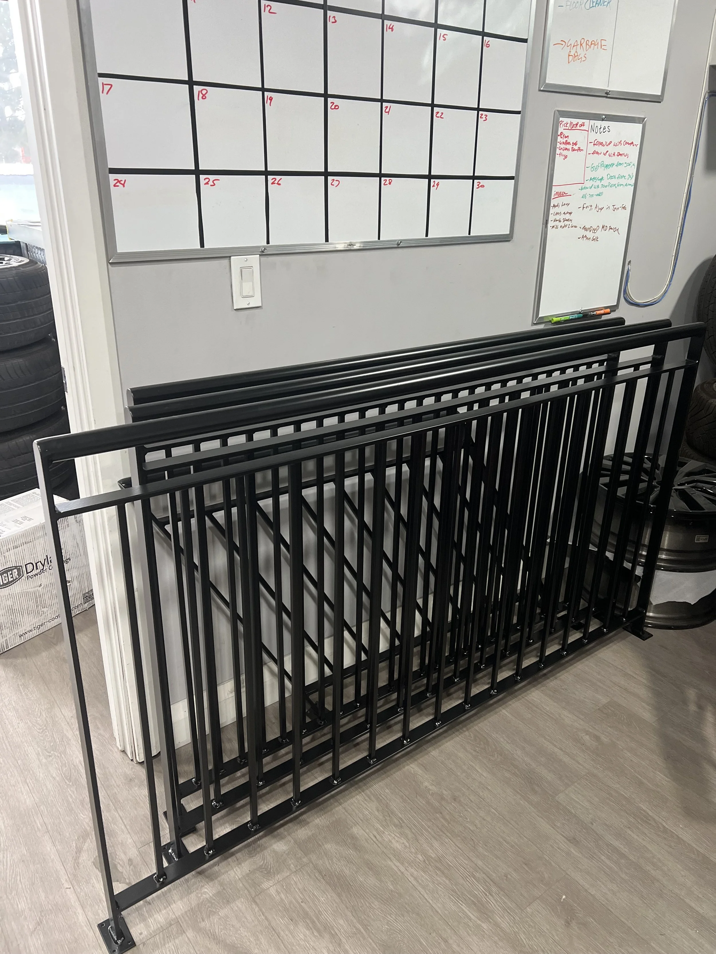 Black metal rack with vertical bars and three horizontal bars across the top, placed indoors against a white wall with a whiteboard and a calendar behind it.